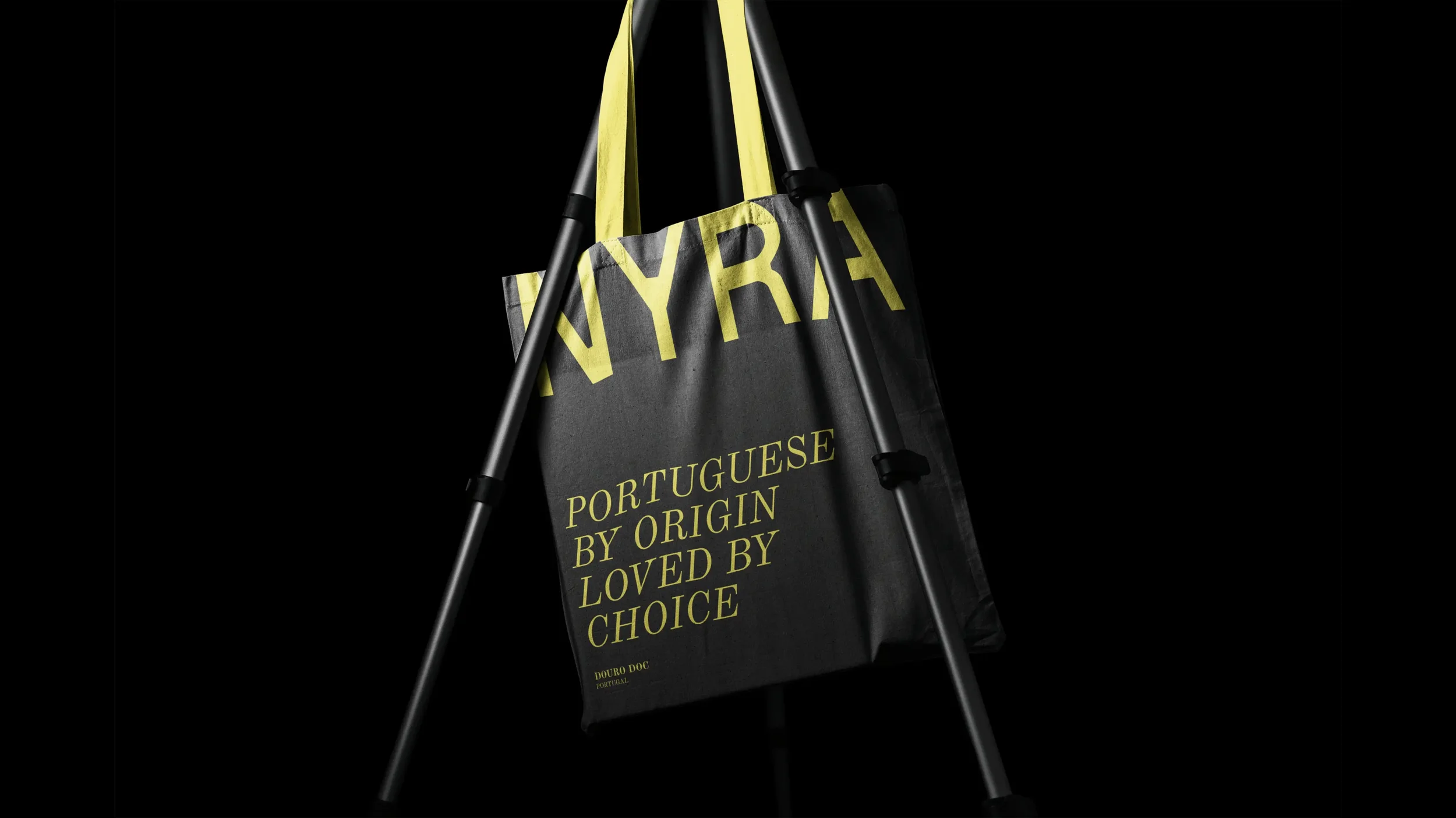 A black tote bag hangs on a metal tripod with yellow straps. The bag has large yellow text that reads 'WYRA' and smaller yellow text that says 'PORTUGUESE BY ORIGIN LOVED BY CHOICE' with 'Douro DOC PORTUGAL' at the bottom.