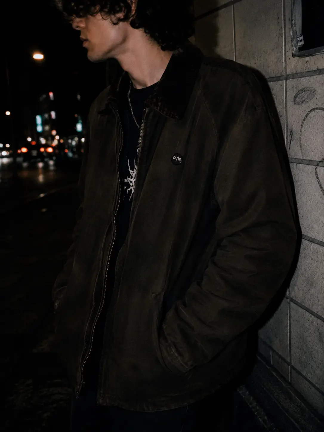 A person with curly hair standing on a city street at night, wearing a dark jacket and chains, with a brick wall behind them and blurred city lights in the distance.