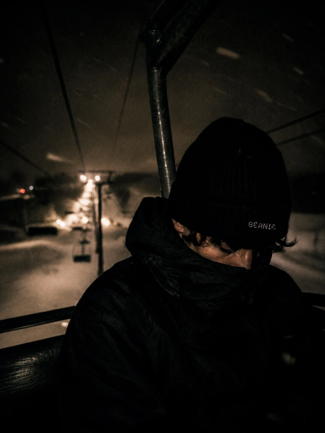 A person wearing a black beanie and jacket is riding an all-terrain vehicle or UTV in a snowy, dark environment at night.