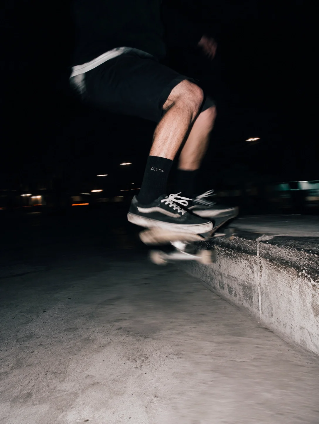 Close-up of a person skateboarding at night, wearing black skateboard shoes, black shorts, and black socks, performing a trick on a concrete ledge.