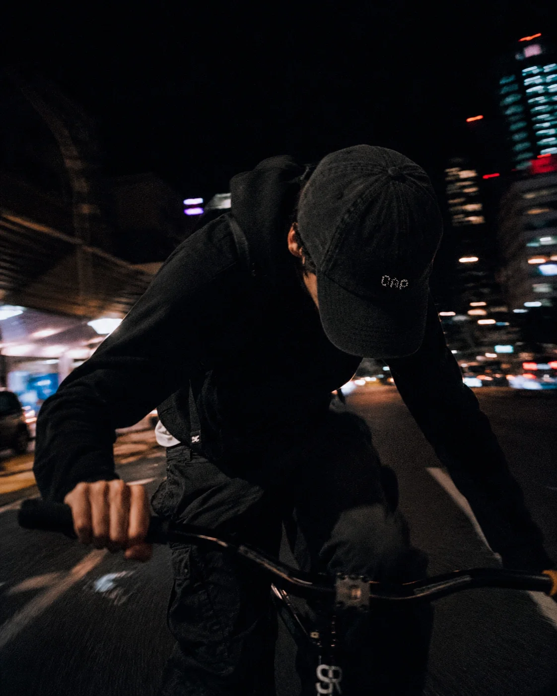Person riding a bicycle at night in an urban area, wearing black clothing and a black cap, with city lights and buildings in the background.