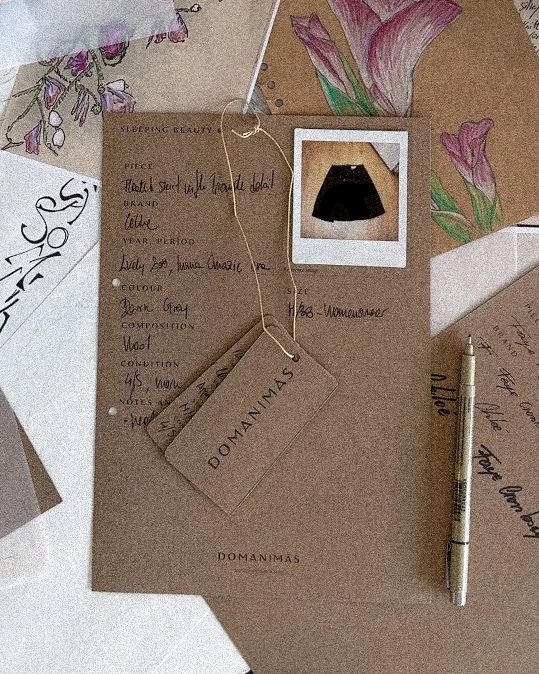A brown paper sheet with handwritten notes and a tag labeled 'DOMANIMAS'. There is a small photograph of a dark-colored skirt, a handwritten note, a beige pen, and decorative floral drawings on the paper. The notes appear to describe a clothing item, likely a skirt.