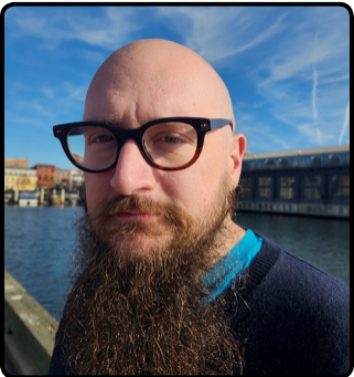 A man with a beard and glasses taking a selfie outdoors near water, with buildings and a blue sky in the background.