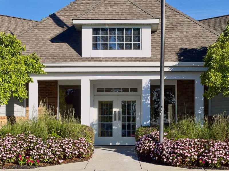 Front of a house with a gabled roof, large front door with glass panels, surrounded by flower beds and trees, and a lamppost in front.