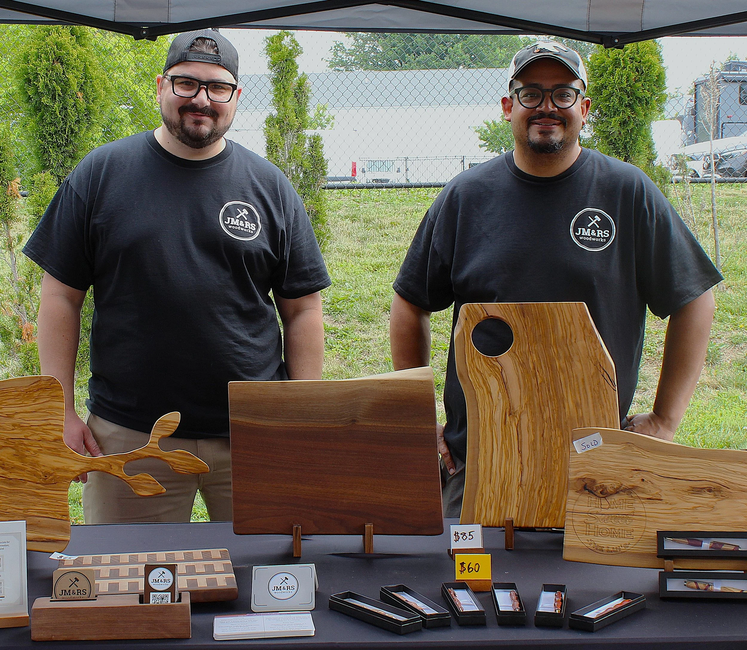 Two men standing behind a display of wooden jewelry and accessories at an outdoor market. The men are wearing black T-shirts and glasses, one with a baseball cap and the other with a cap. Behind them is a chain-link fence with trees and vehicles in the background.