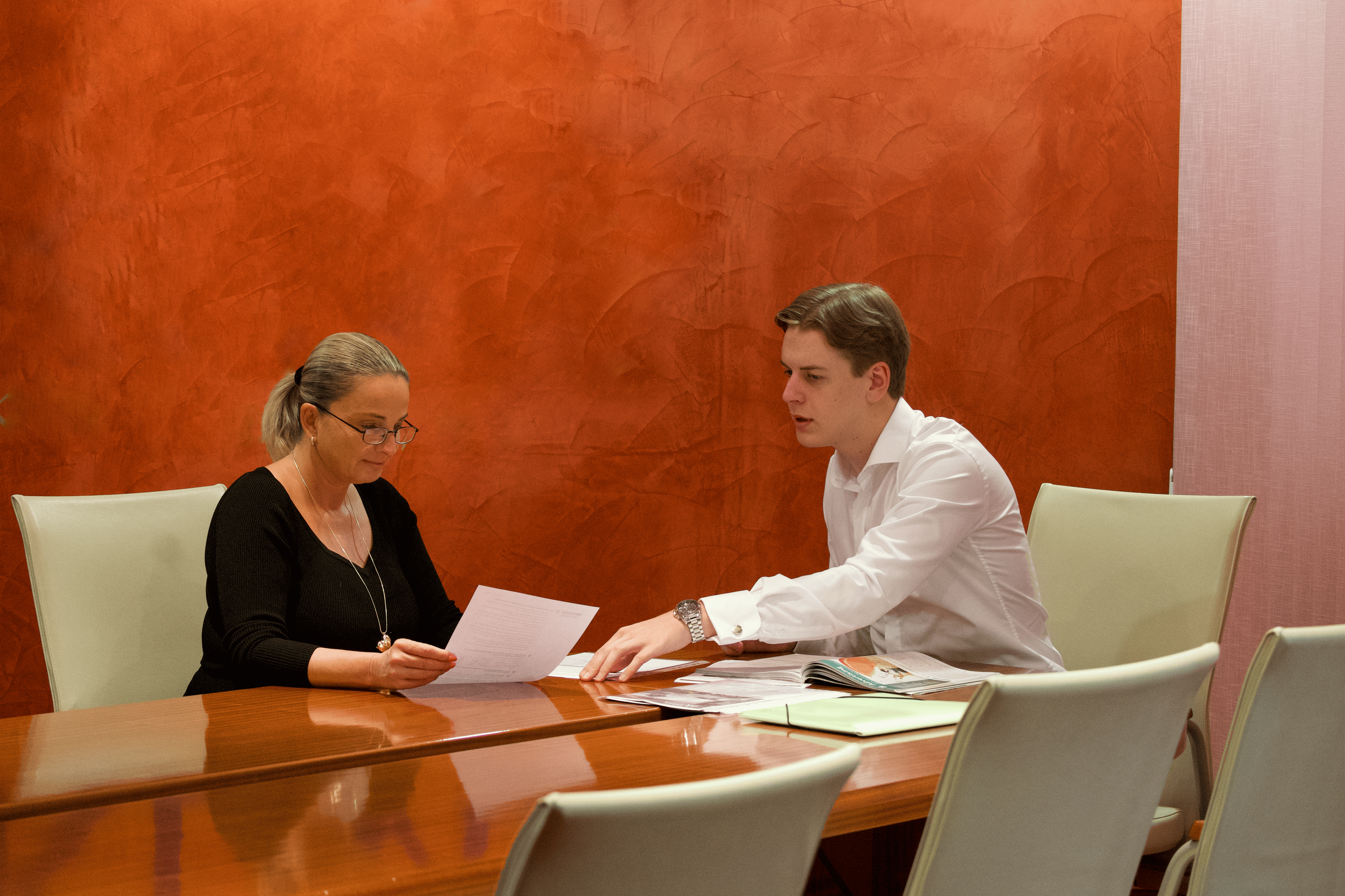 A business english tutor and acorporate student sit at a conference table reviewing documents in a meeting room, studying business english