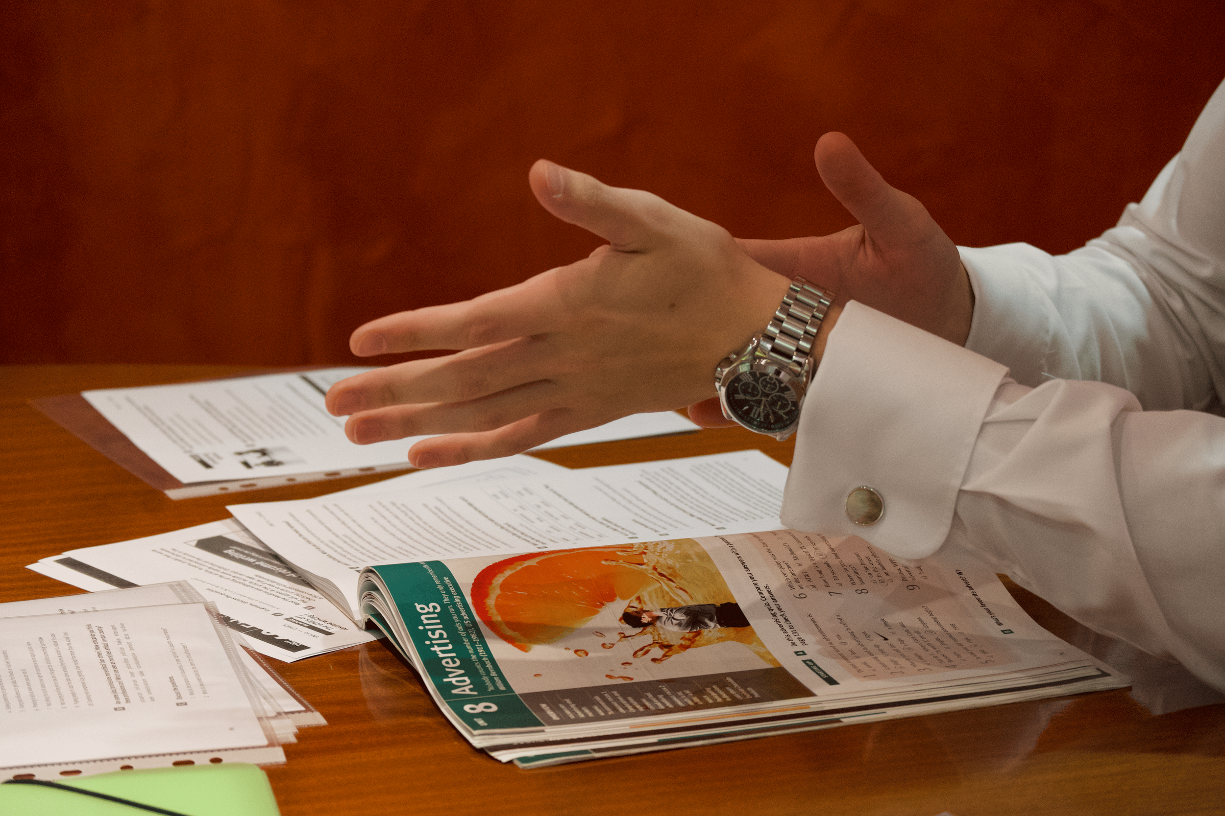 A Business English tutor in a white shirt with cufflinks and a silver watch gesturing over a table with Business English materials and an open coursebook.