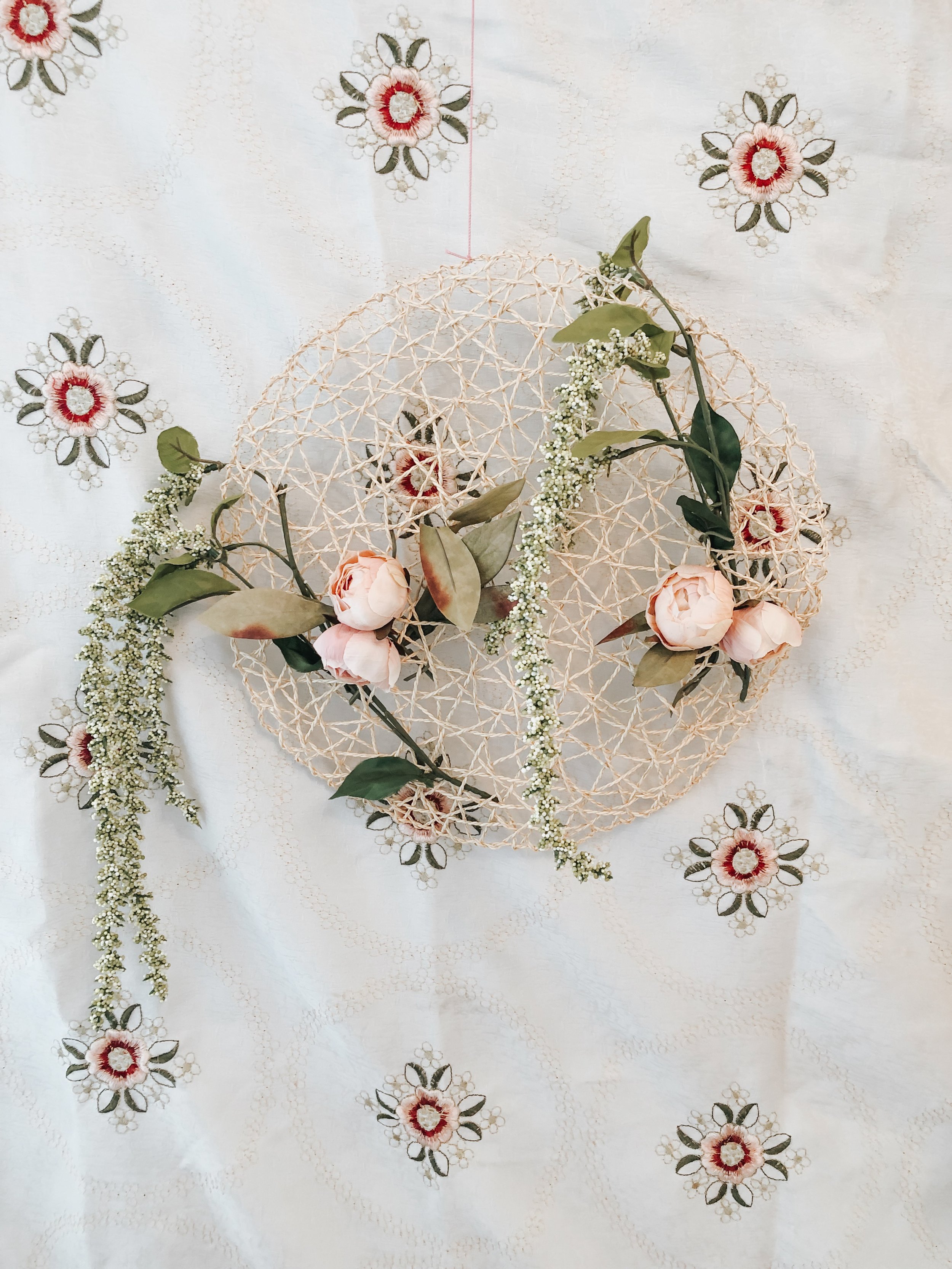 Decorative floral arrangement with pink roses, green leaves, and white small flowers on a beige woven spherical frame, placed on embroidered fabric with floral patterns.