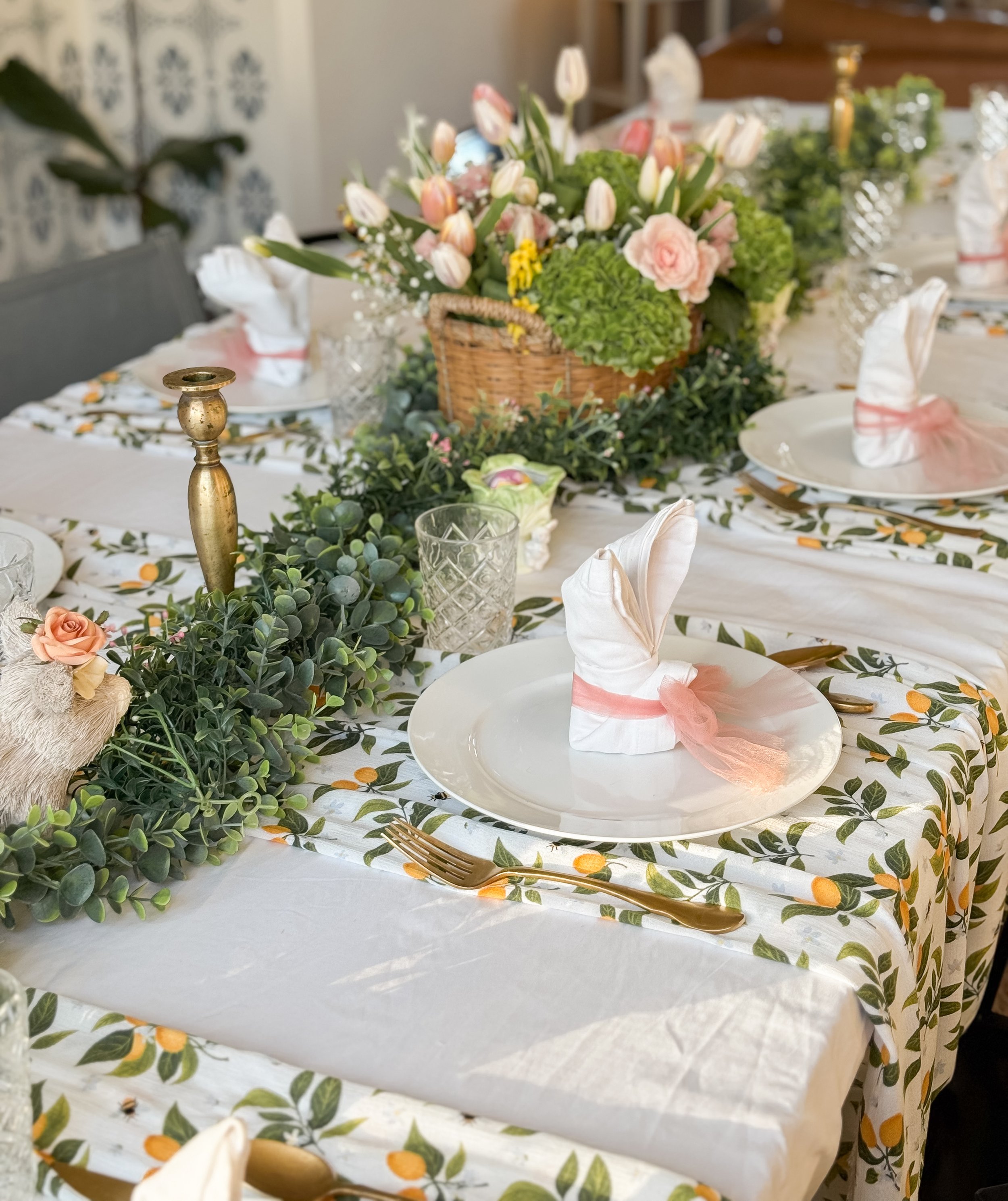 Decorated dining table with white tablecloth featuring a fruit and leaf pattern, set with white plates, gold cutlery, crystal glasses, and napkins folded with pink ribbon. Centerpiece includes a lush green garland, a basket of pink and white tulips a
