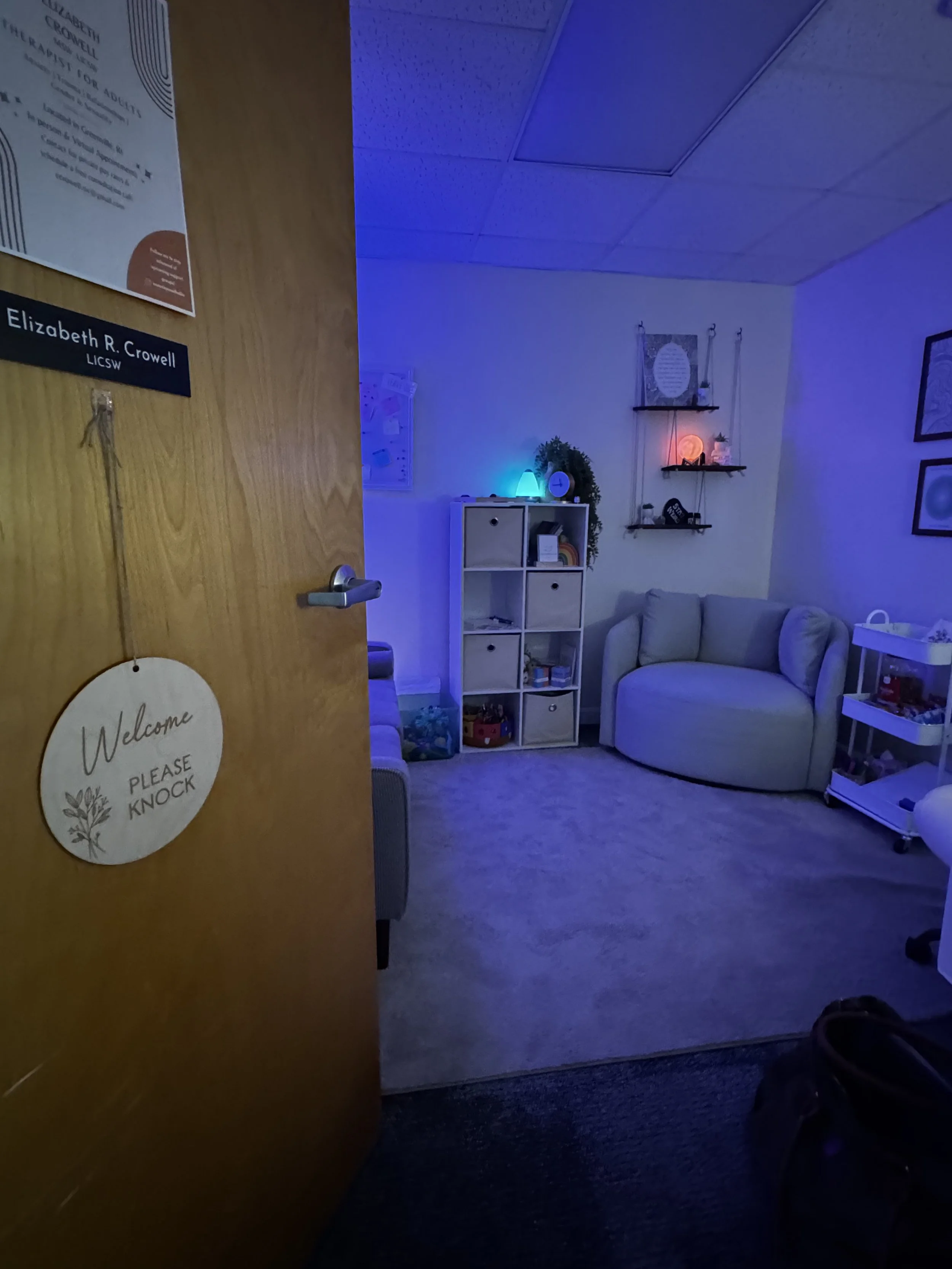 A cozy therapy room with a beige couch, white shelving unit, and wall decor, illuminated by blue and warm lighting.