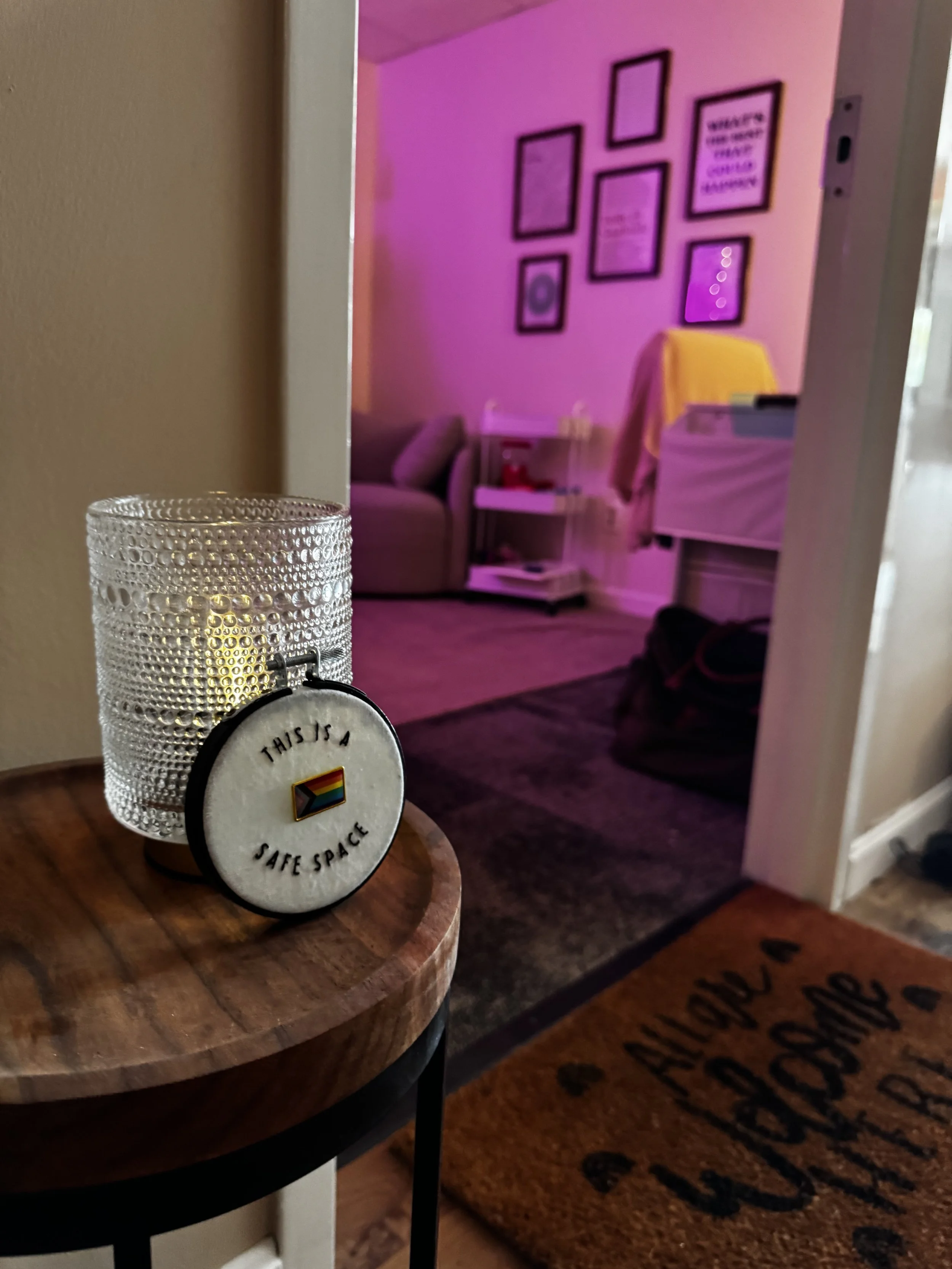 Decorative candle holder and a circular pin with a pride flag and the words "This is a safe space" in a cozy room with purple lighting, containing a sofa, framed pictures on the wall, and various furnishings.