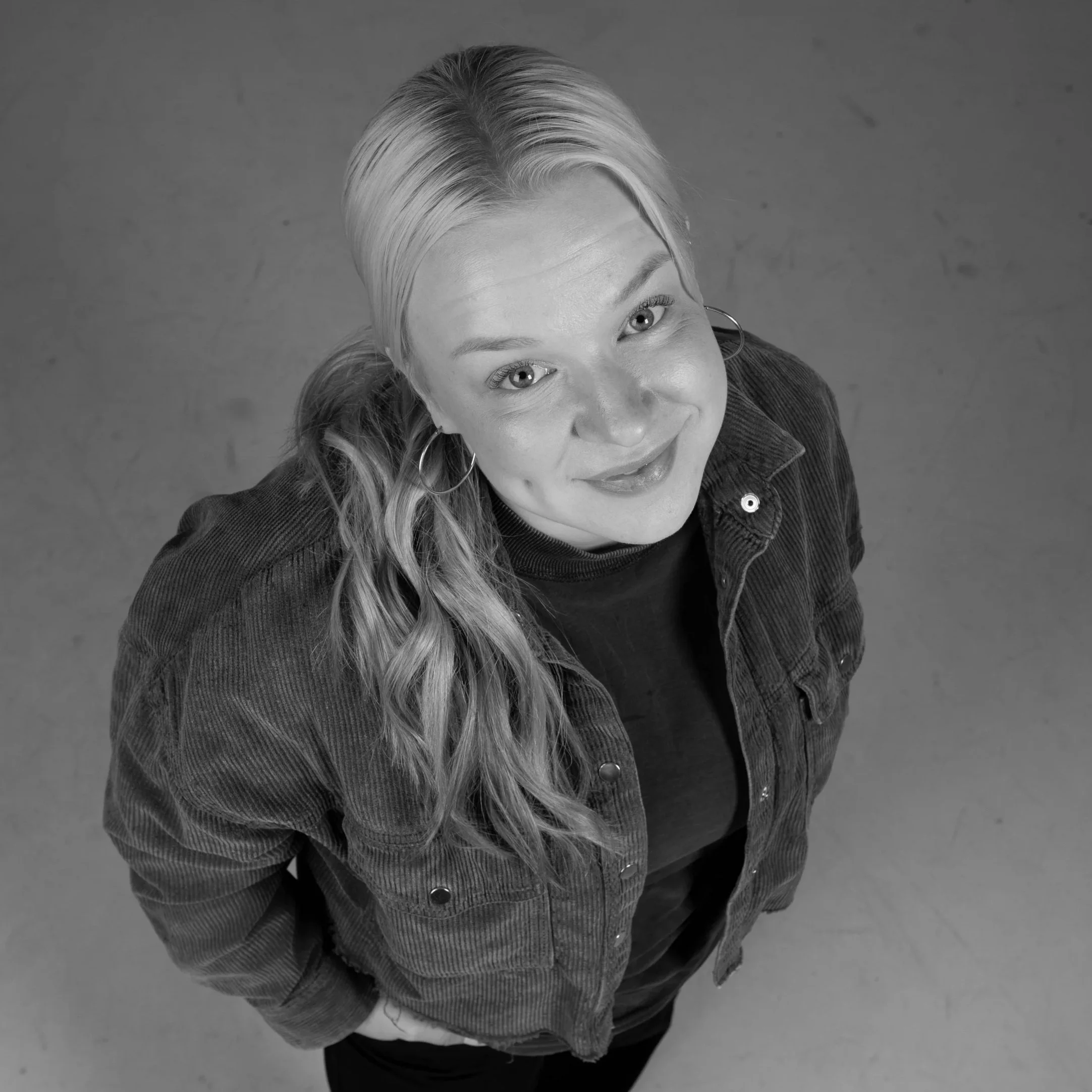 Väritön kuva nuoresta naisesta, jolla on vaaleat hiukset ja isot korvakorut, katsoen ylöspäin, taustalla studio.