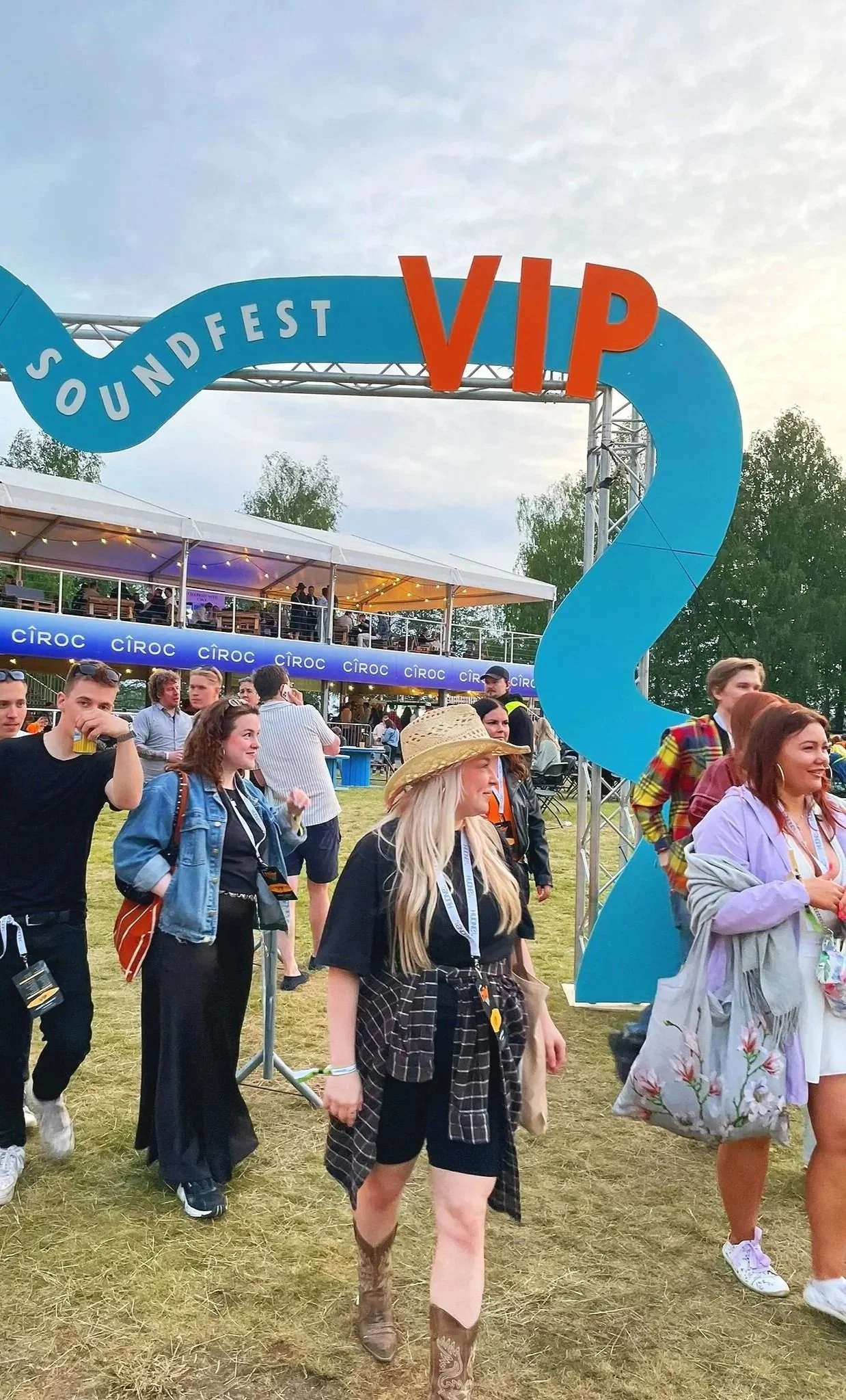 Ihmisiä festivaalialueella, taustalla suuri sininen ja oranssi VIP-alueen merkki, joissa lukee 'Soundfest VIP', ja ympärillä puistomainen ympäristö.