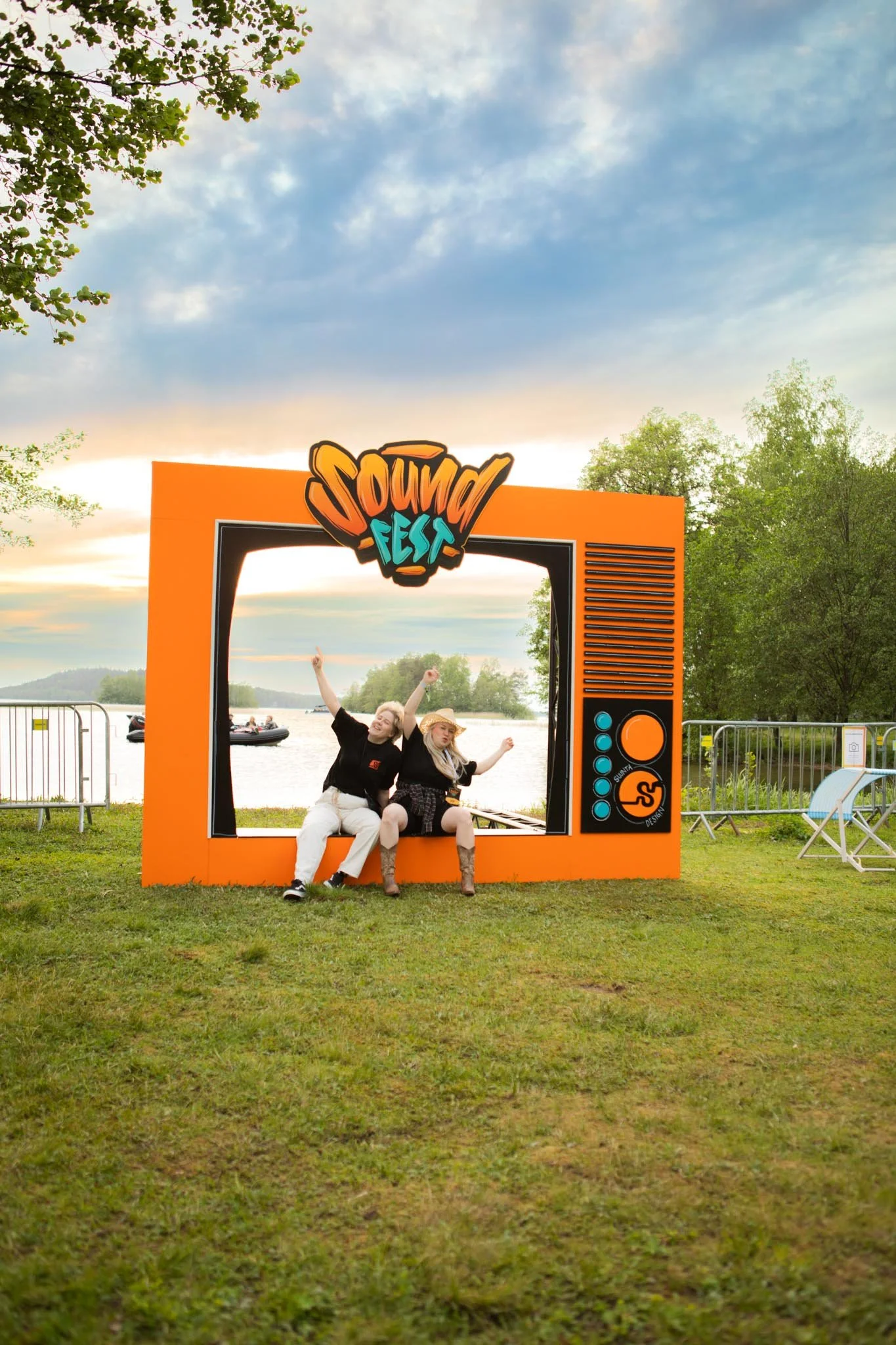 Kaksi ihmistä istuu valokuvausseinassa, joka on muotoiltu vanhan television muotoiseksi ja jossa lukee 'Sound Fest'. Taustalla järvi ja puissa vihreitä lehtiä, taivaalla pilviä ja auringonlasku.