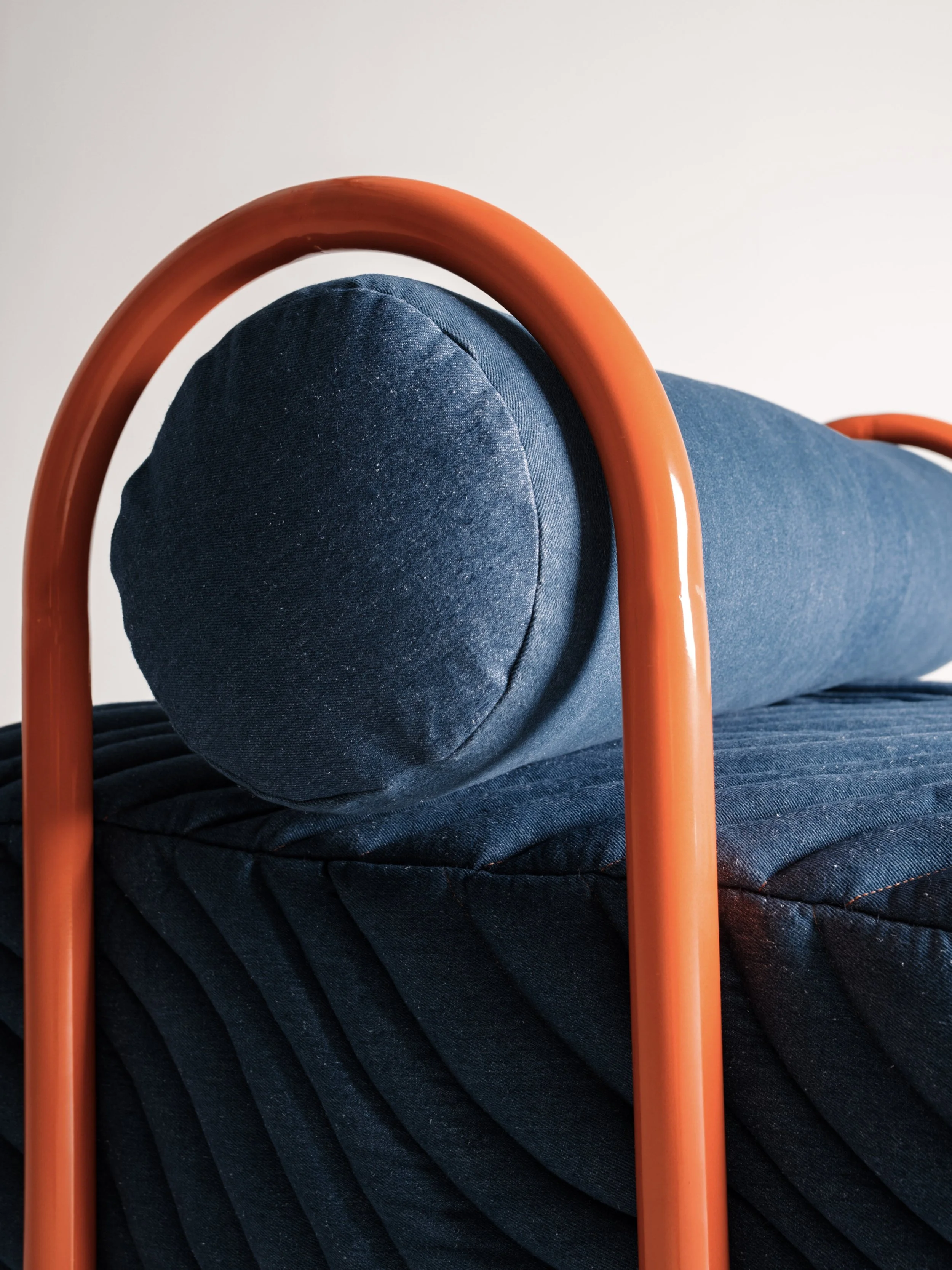 Välissä näkyy sininen tyyny, jonka päällinen on tehty farkku-materiaalista, ja se on asetettu sohvan päälle. Sohvassa on myös tummansininen pehmuste, ja taustalla on valkoinen seinä. Kuvassa on myös kaksi oranssia metalliputkea, jotka muodostavat osan huonekalusta.