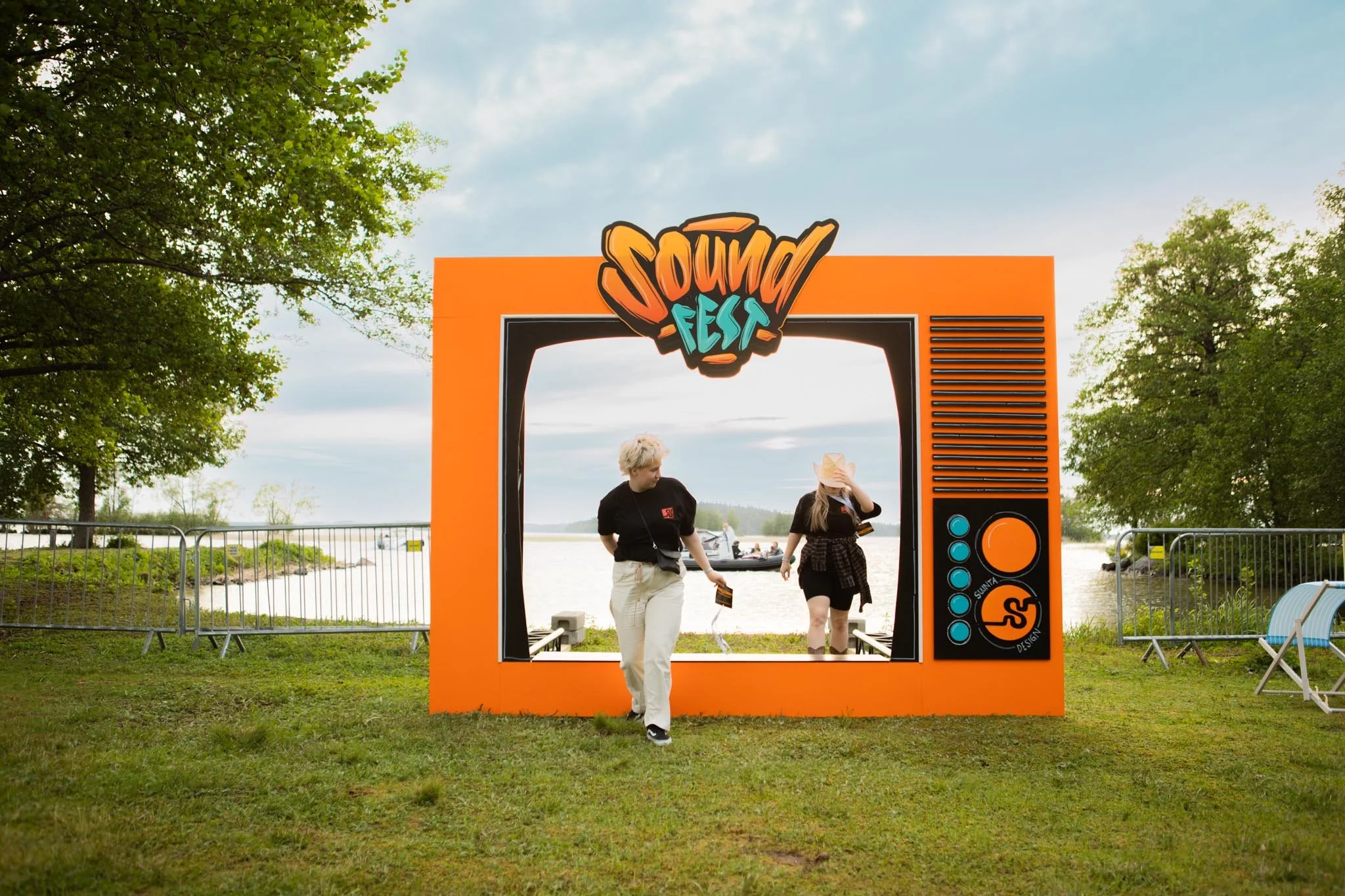 Kaksi muotoilijaa kävelee läpi suuresta, oranssista Soundfestin kuvausportista, jossa on grafiikkamaalaus. Taustalla on järvi ja vihreitä puita, taivas on pilvinen.