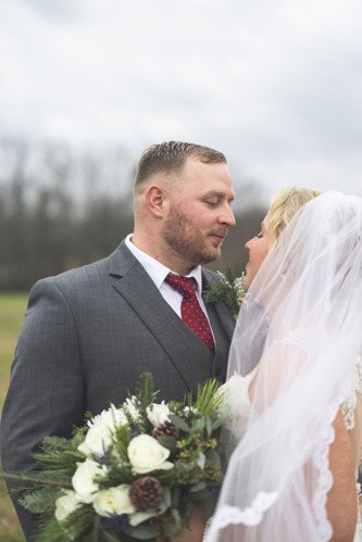 Bride and groom gazing at each other outdoors, bride holding a bouquet of white flowers and greenery, wedding attire, overcast sky.