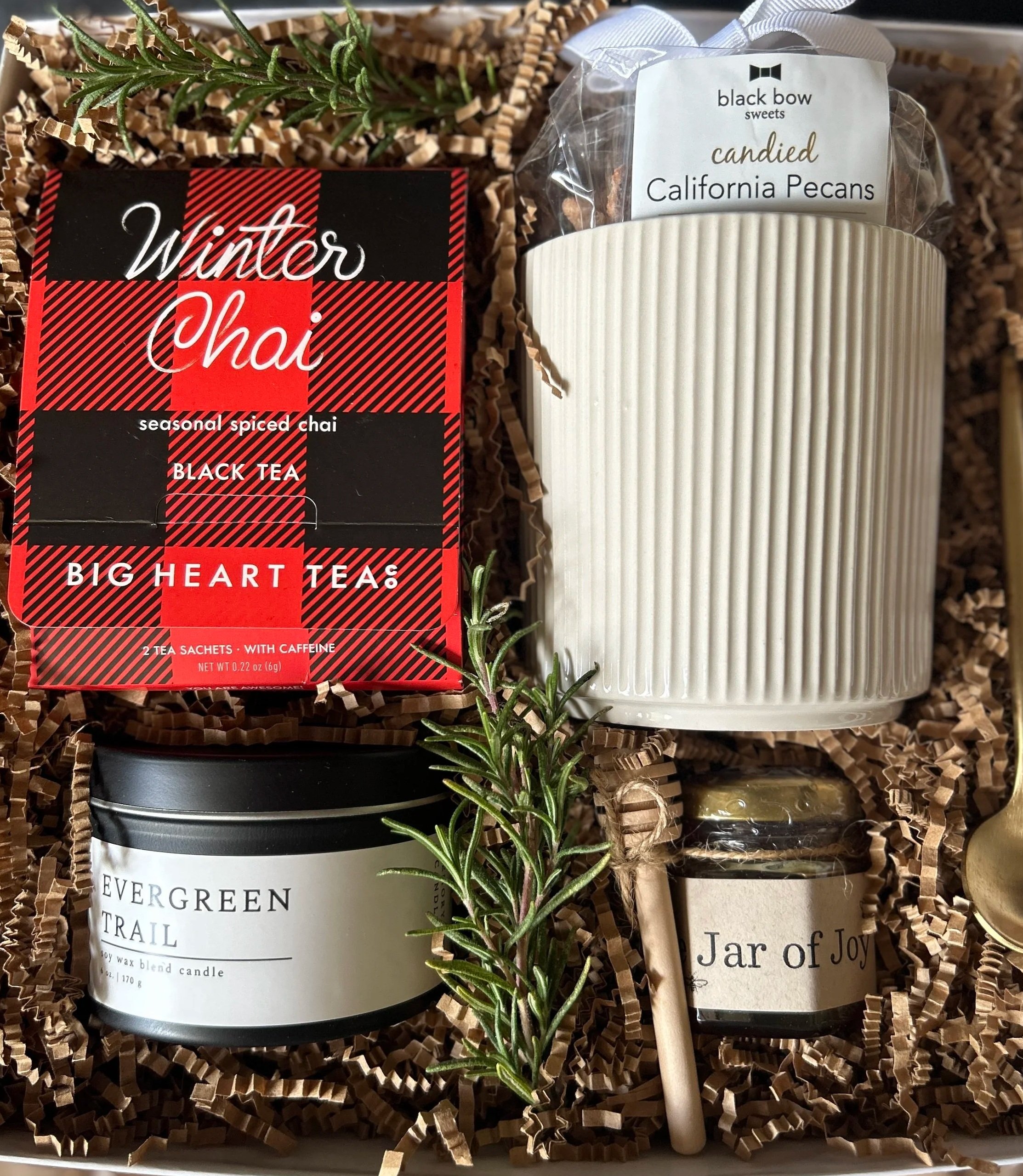 A gift box contains holuday tea, an evergreen trail candle jar of wildflower honey and mug and california candied pecans with a gold tea spoon christmas greens and neutral crinkle paper