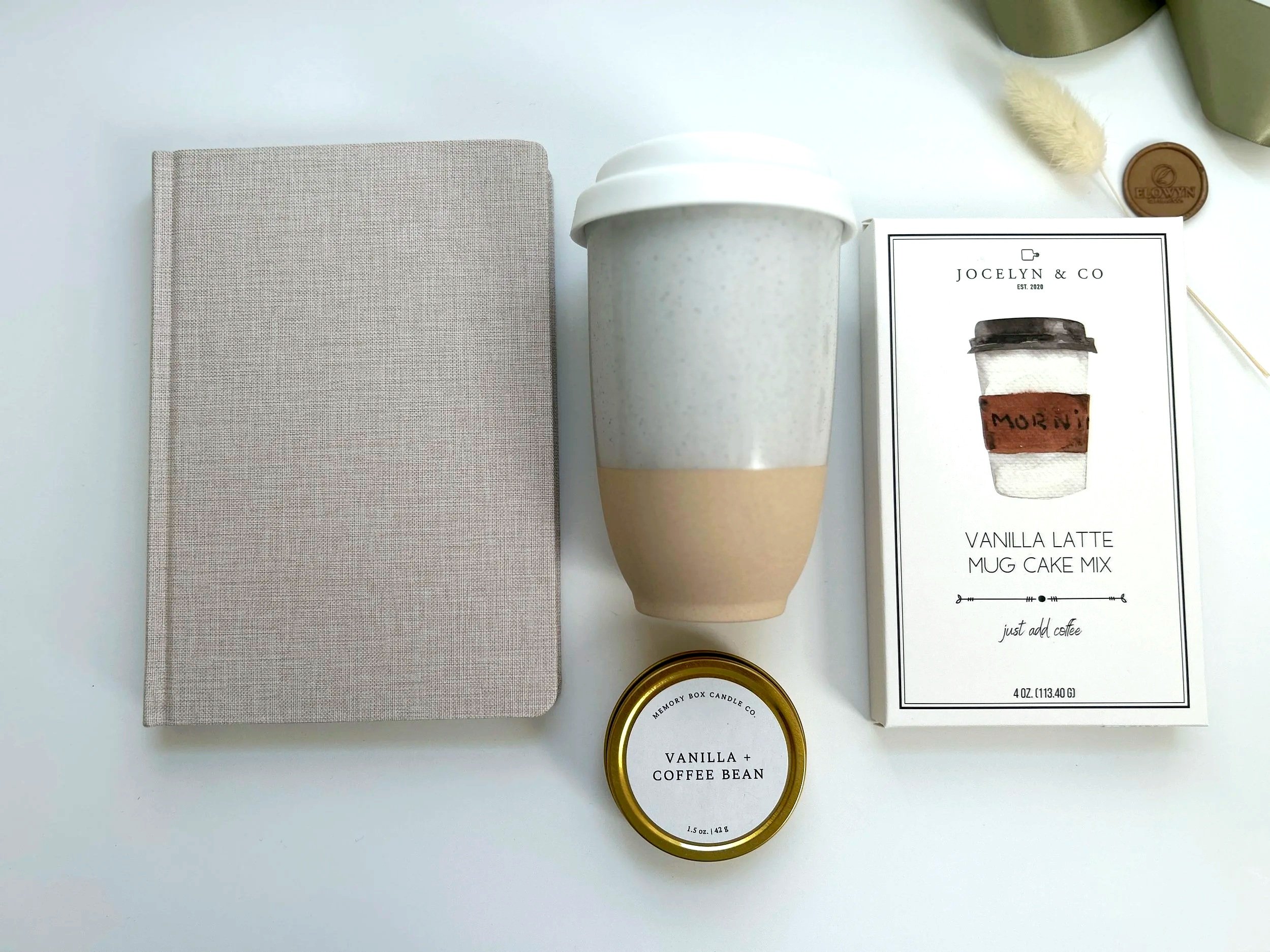 Gift box containing lavender eye pillow, ceramic tumbler, lavender and rosemary essential oil, and herbal tea bag, all arranged with shredded paper and burlap background.