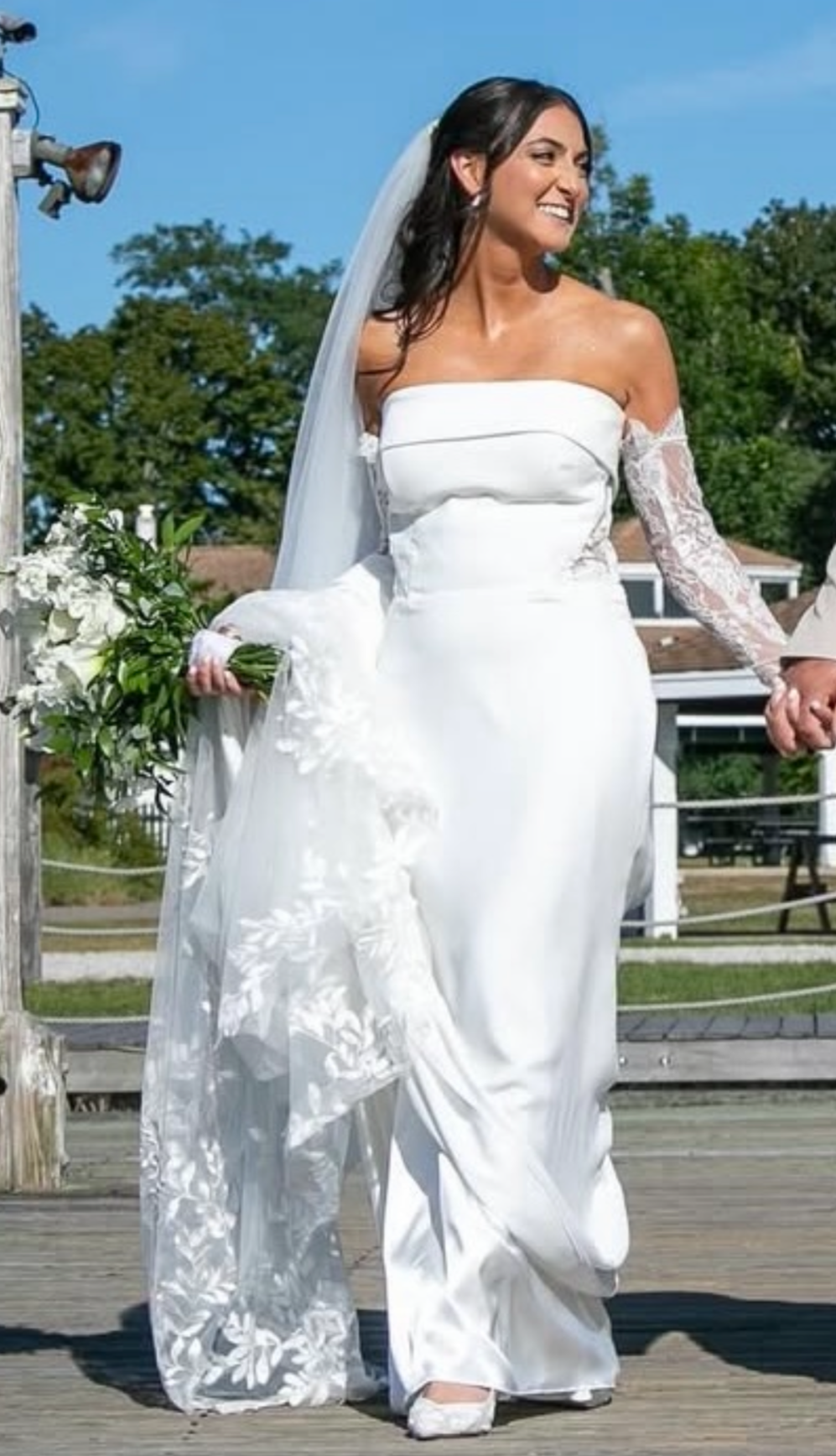 A bride in a white wedding gown holding a bouquet of white flowers, standing outdoors on a wooden deck, smiling.