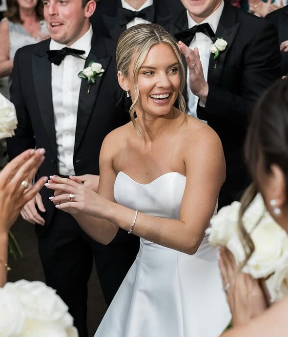 A bride in a white strapless dress smiling and clapping, surrounded by groomsmen in black tuxedos with white shirts and black bow ties at a wedding reception.