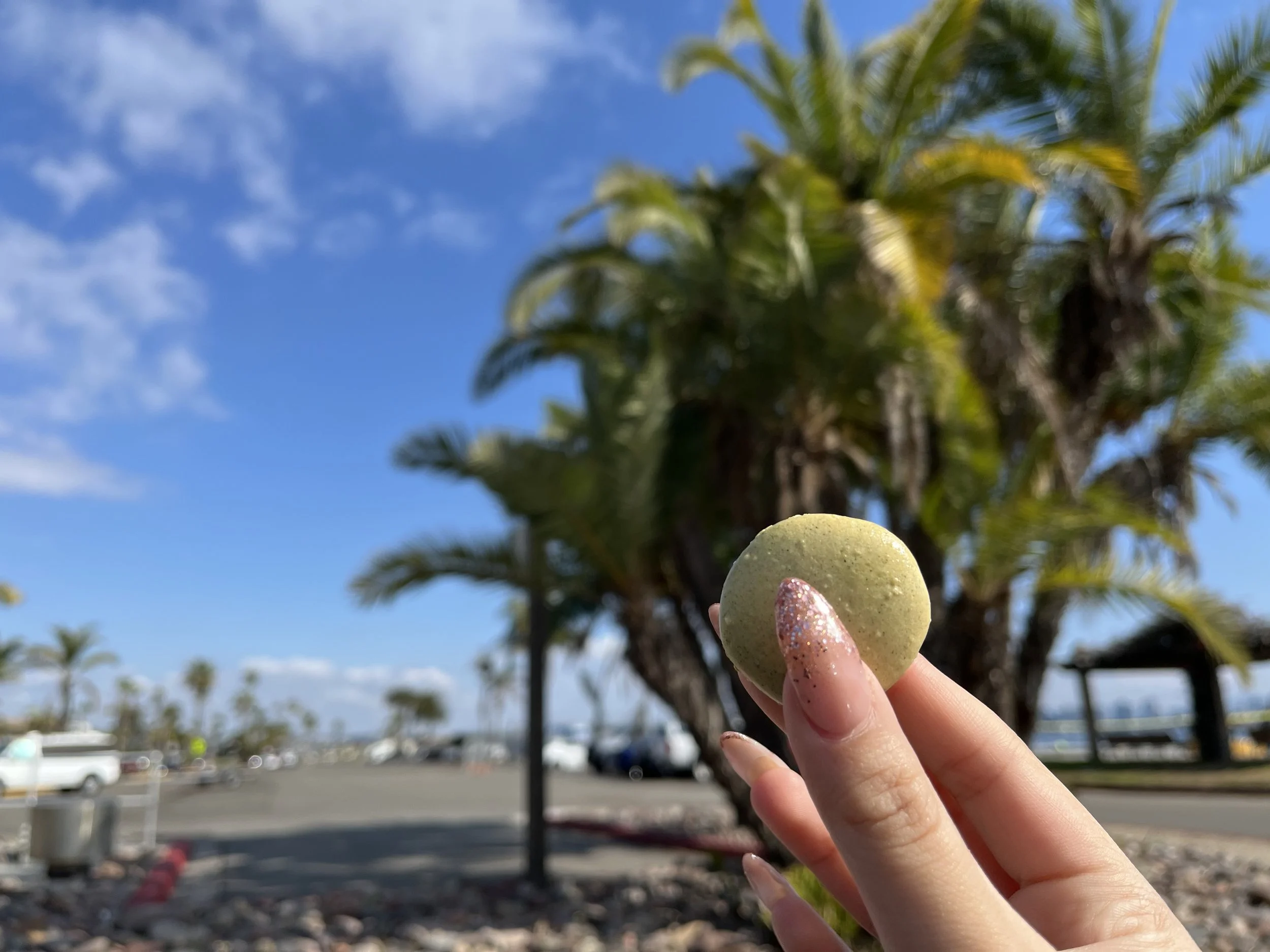 Hand holding a green macaron with glitter against a backdrop of palm trees, blue sky, and parking lot.