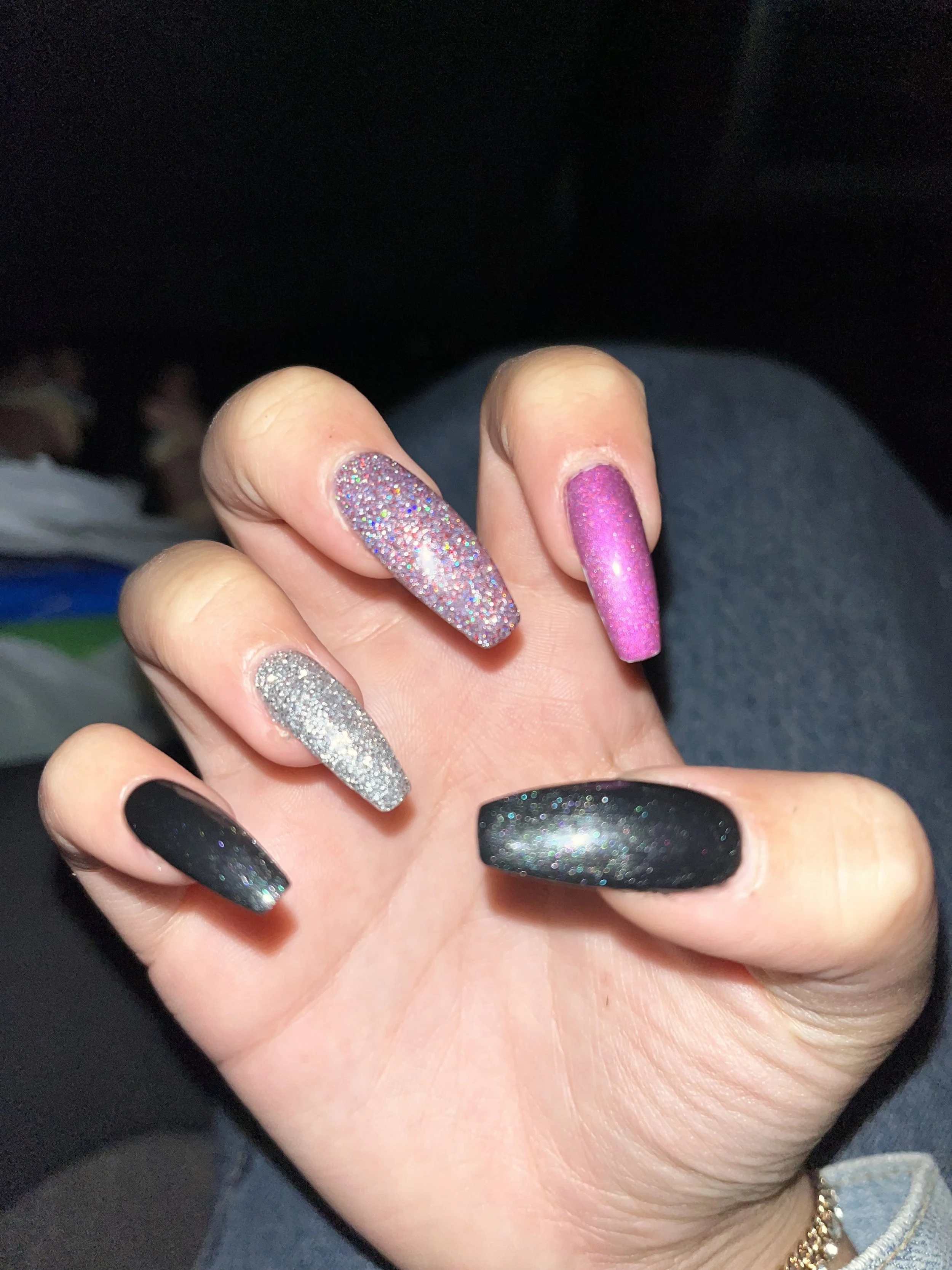 A hand with five decorated nails featuring various glittery and matte nail polish designs in colors silver, pink, black, and multicolor glitter.