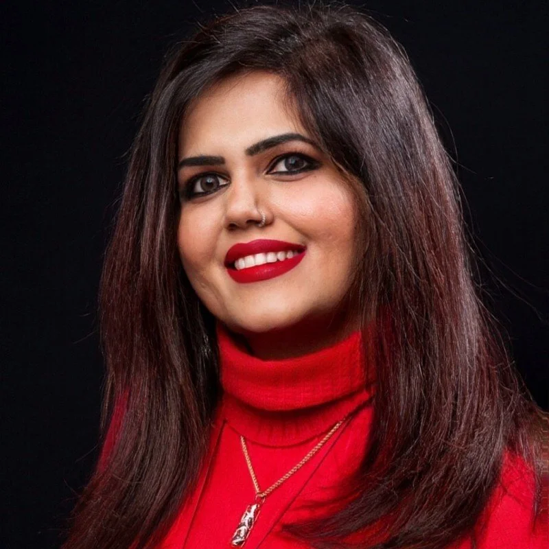 A woman with dark brown hair, wearing a red turtleneck and gold jewelry, smiling against a black background.