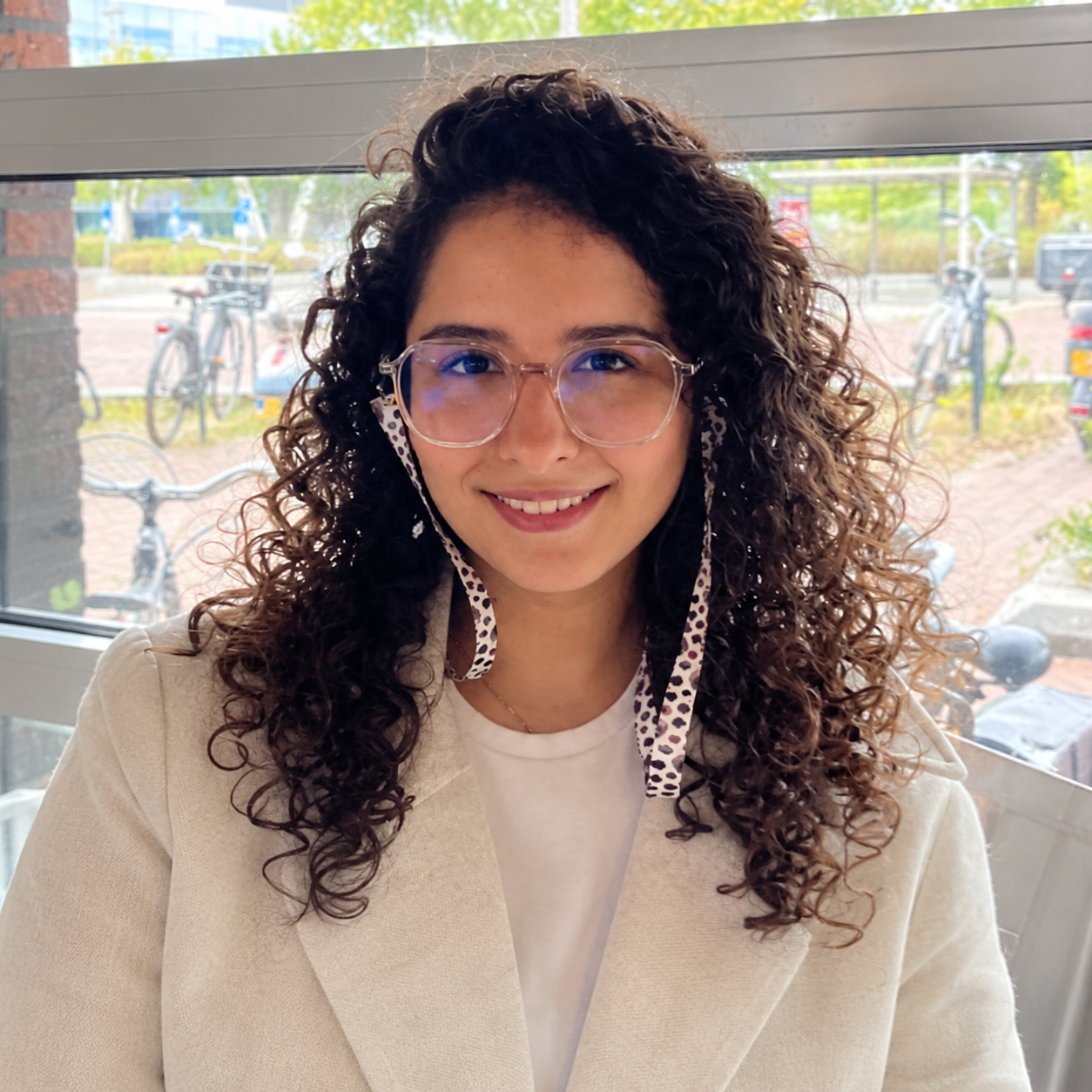 A woman with curly hair and glasses smiling at the camera, sitting indoors near a window with bicycles outside.