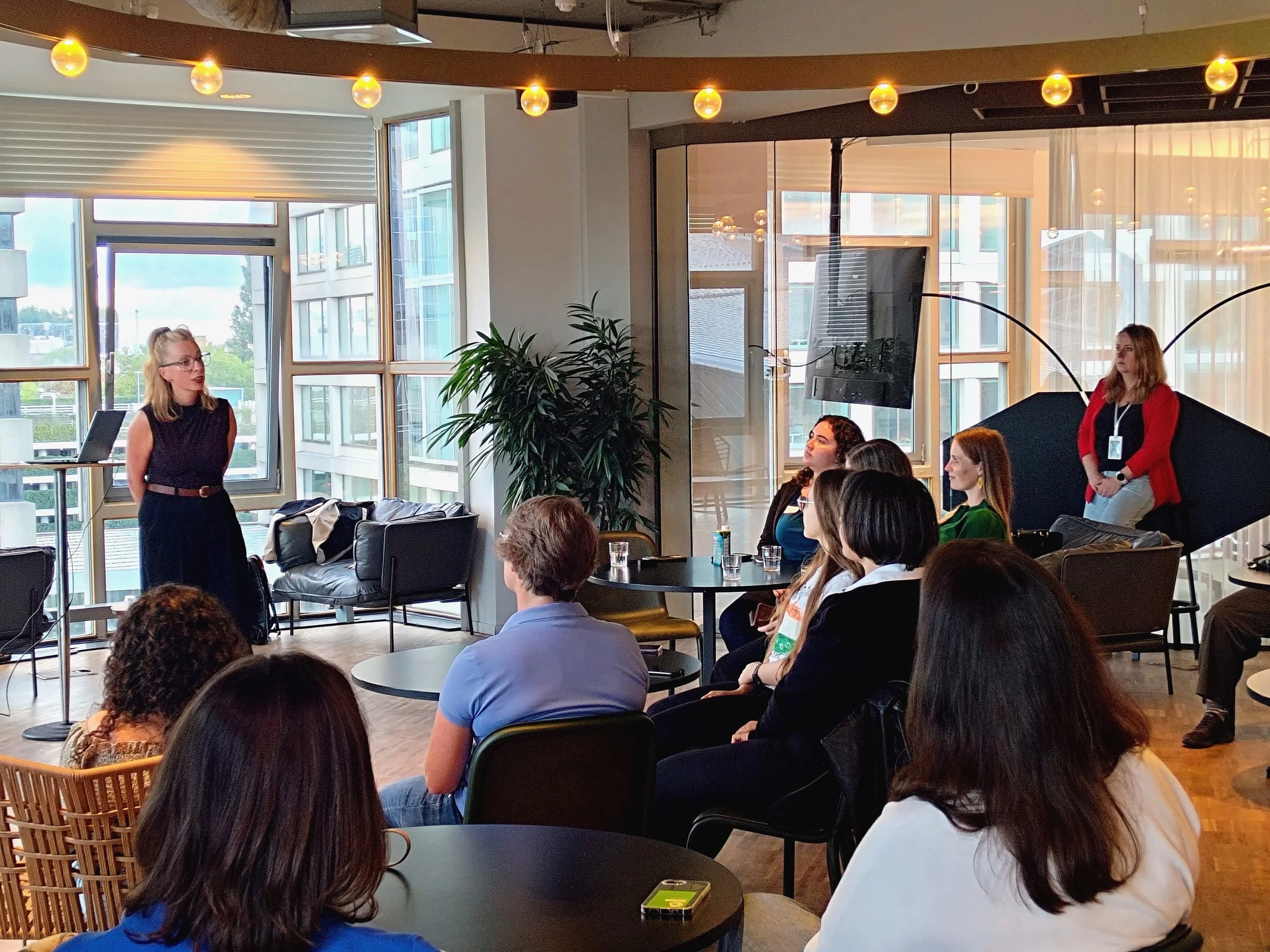 Group of women seated at tables listening to a woman standing and speaking in a modern, well-lit conference room with large windows and indoor plants.