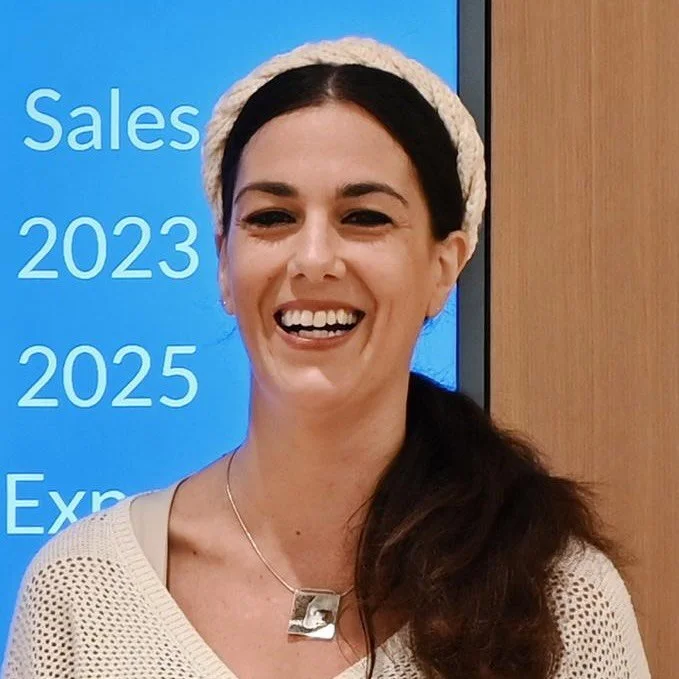 A cheerful woman with dark hair wearing a cream-colored knit hat and necklace, standing in front of a blue screen displaying text about sales for 2023 and 2025.