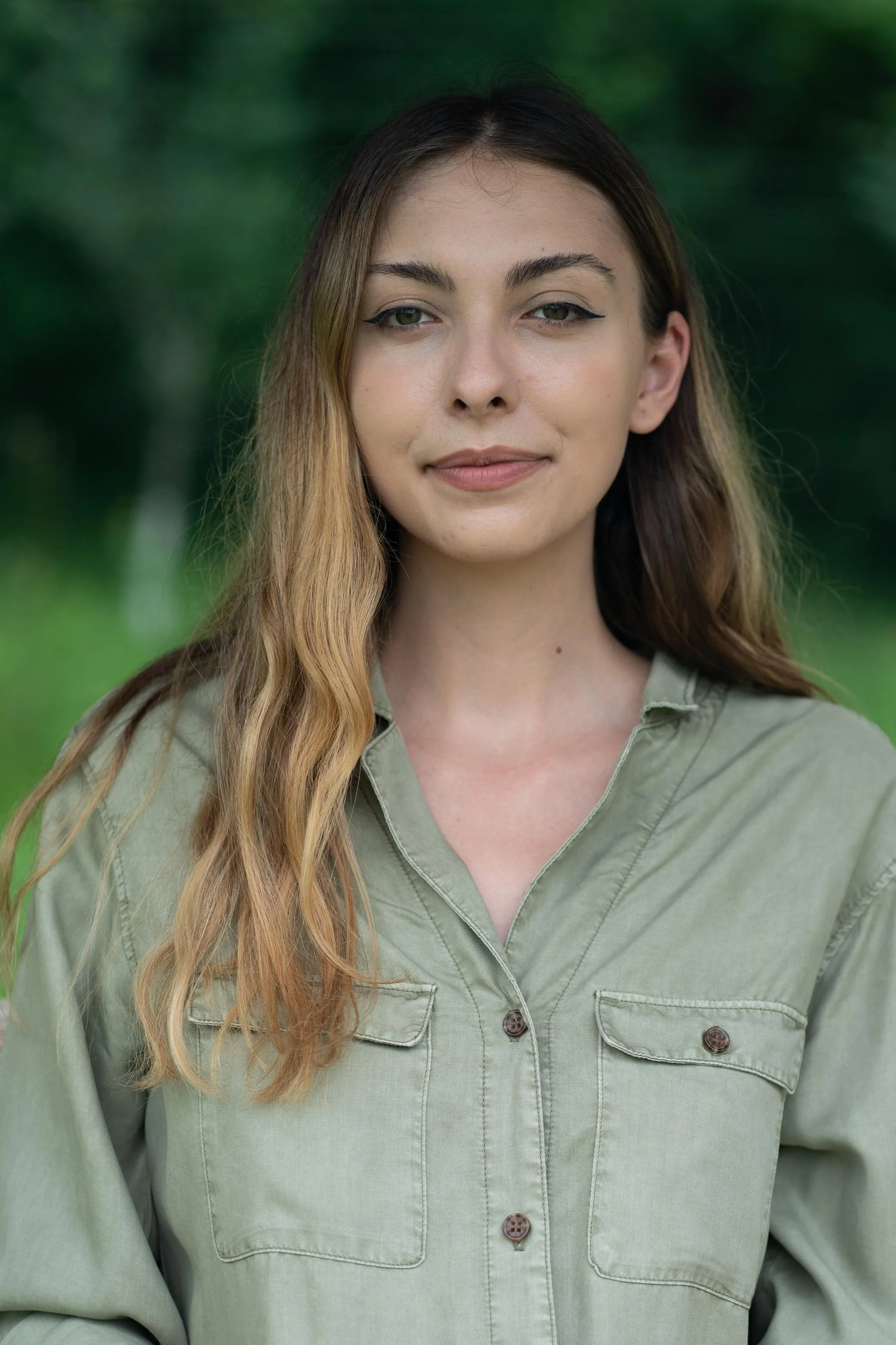 A young woman wearing a light green shirt outdoors with a blurred green background, looking at the camera with a slight smile.