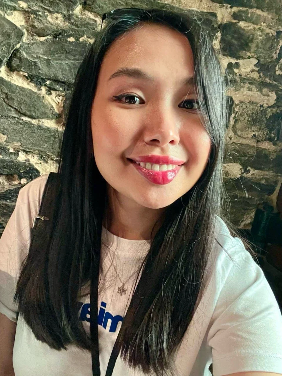 A woman with long black hair smiling at the camera, wearing a white shirt and a small necklace, with a stone wall background.