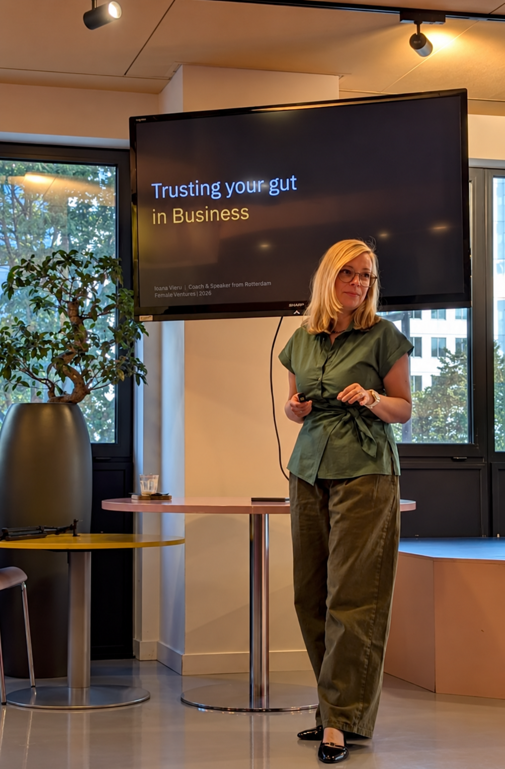 A woman giving a presentation in a modern office space, with a large screen behind her displaying the presentation title "Trusting your gut in Business." The woman has blonde hair, wears glasses, a green shirt, and olive-green pants, and holds a remote control in her hand.