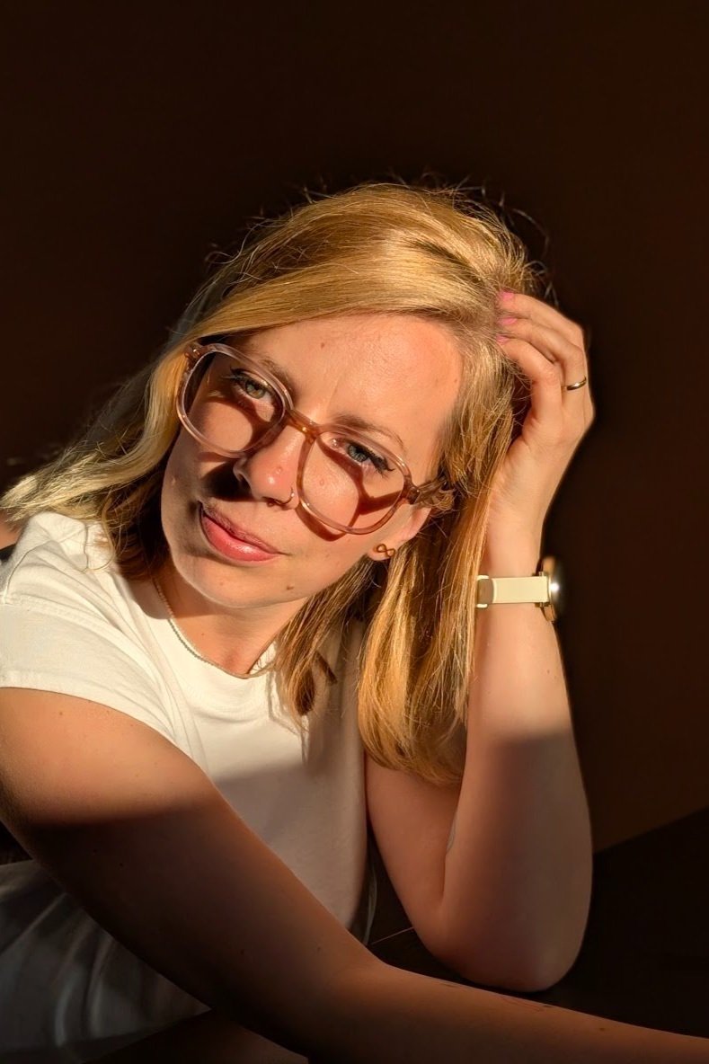A woman with blonde hair and glasses, wearing a white shirt, sitting in sunlight with her hand on her hair, looking at the camera.