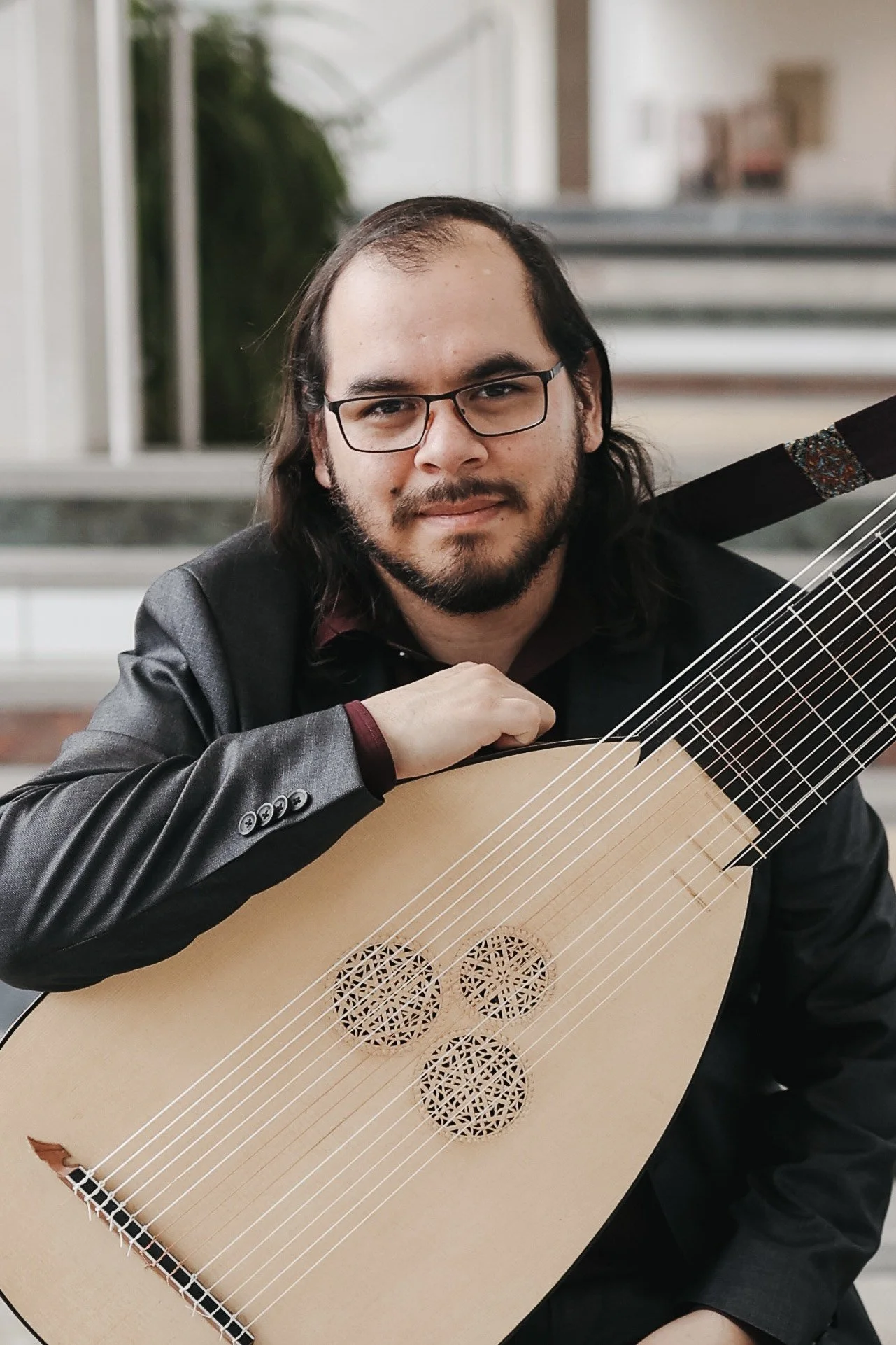 A man with glasses and long dark hair holding a lute, looking at the camera. He is wearing a black blazer and is sitting in a modern indoor setting.