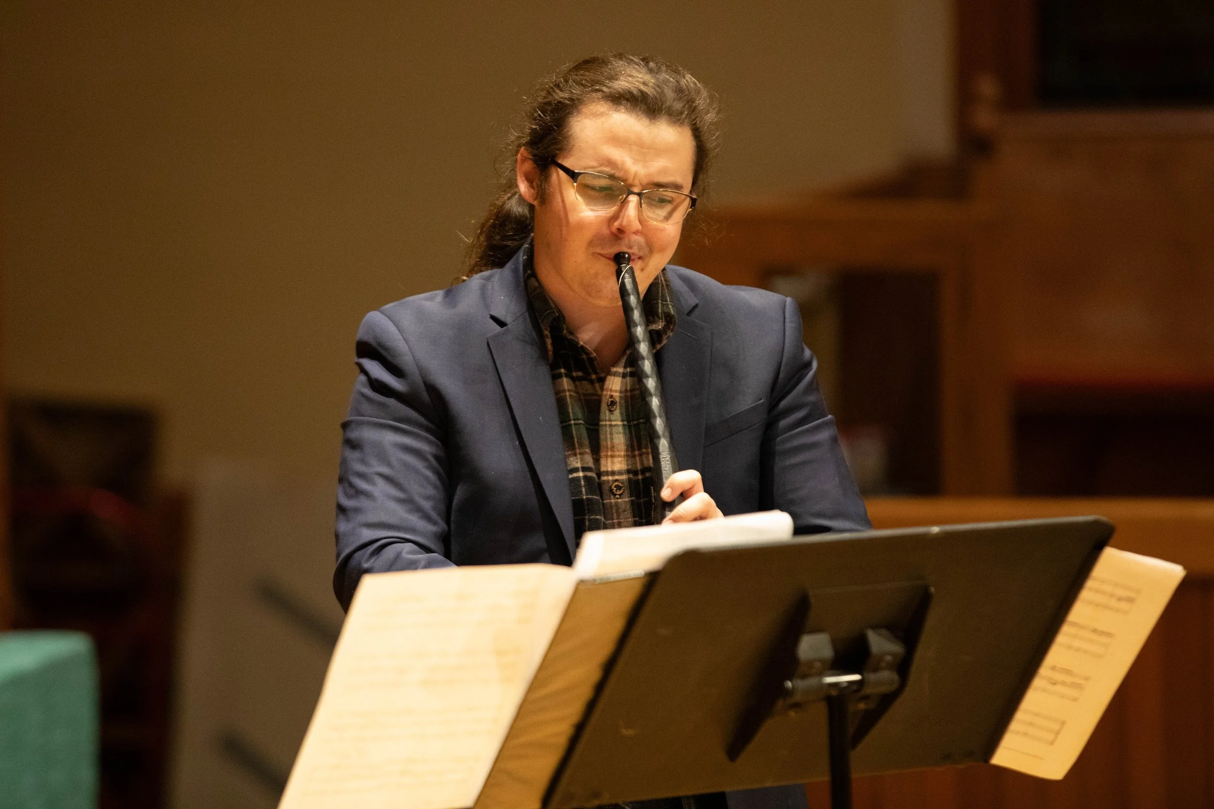 A man with glasses and long hair playing a cornetto while reading sheet music.