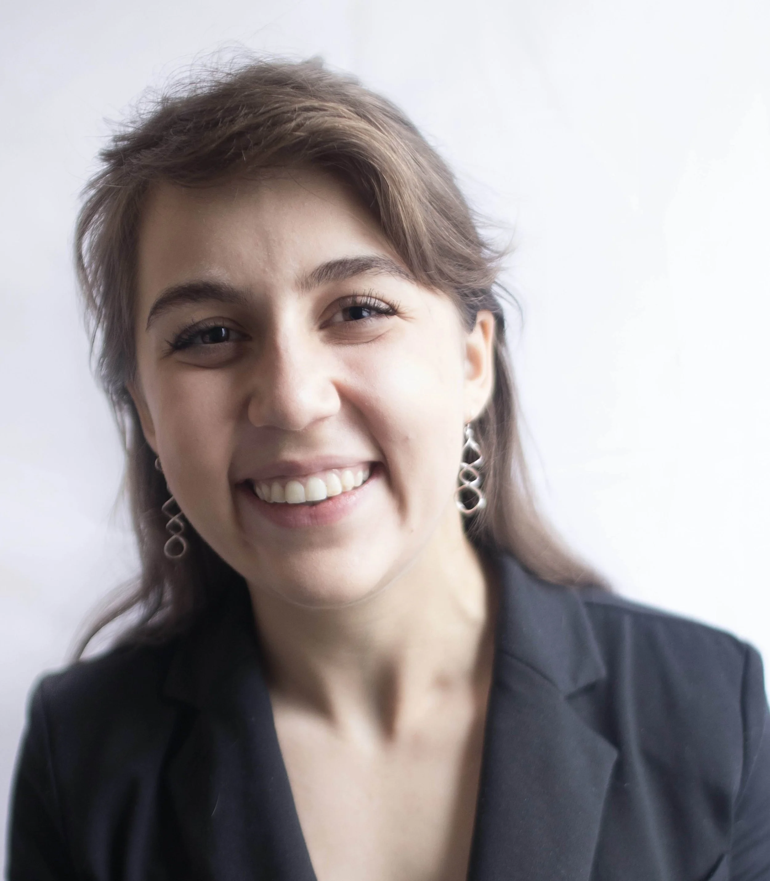 Portrait of a young woman with light brown hair, smiling, wearing a black blazer and silver earrings, against a plain light background.