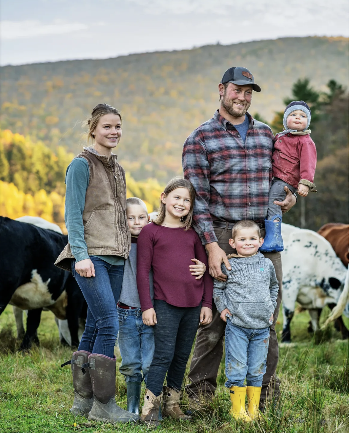 The Gateway Farm Family, including two adults and four children, standing outdoors on a farm with cows grazing nearby and a mountainous landscape in the background. They are dressed in casual farm attire, with some children wearing rain boots.