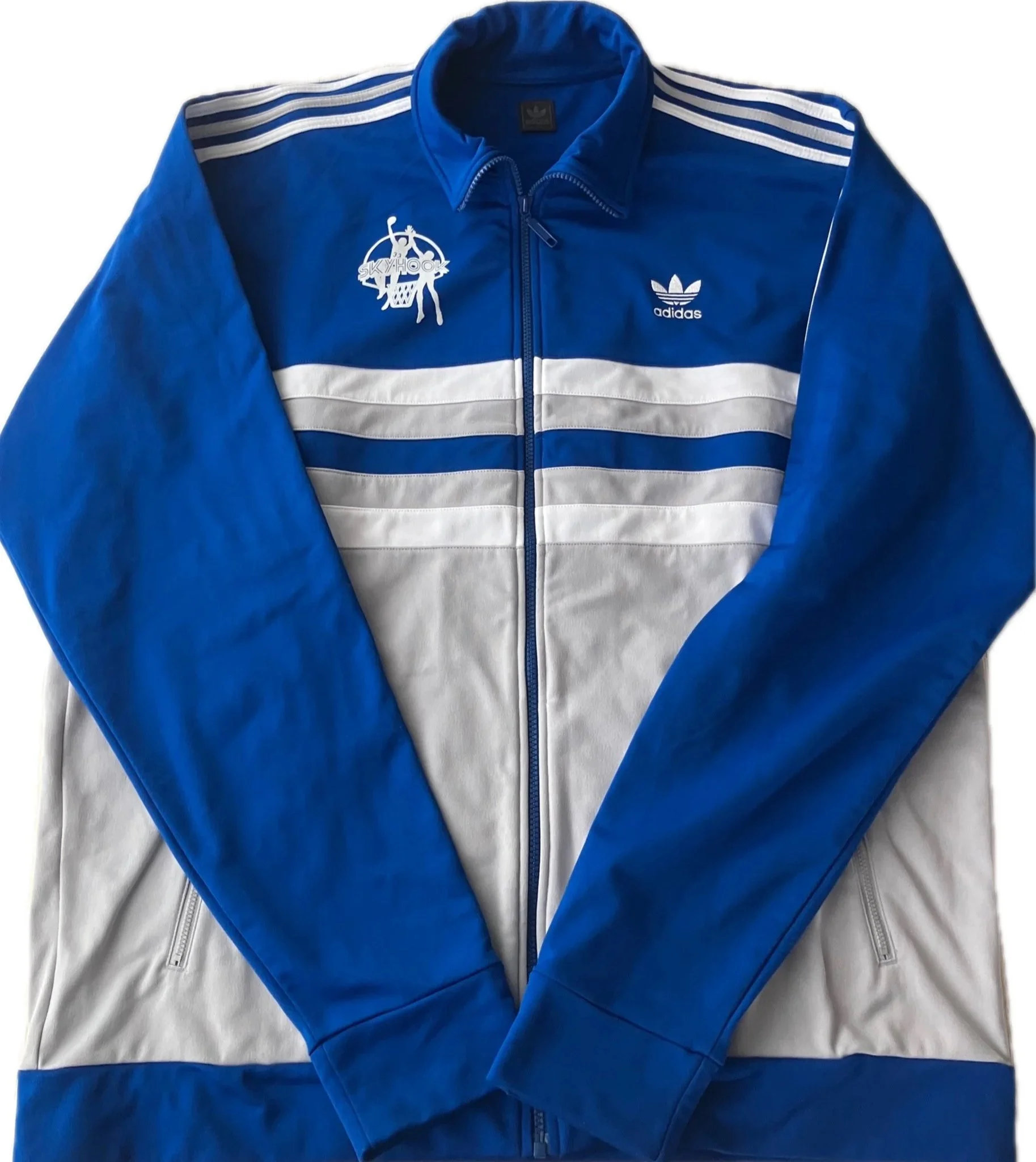 Blue Adidas windbreaker jacket with white stripes on the shoulders, featuring the Adidas trefoil logo and a design with a basketball player silhouette and the word "Skyhook" on the left chest area, laid flat on a white surface.