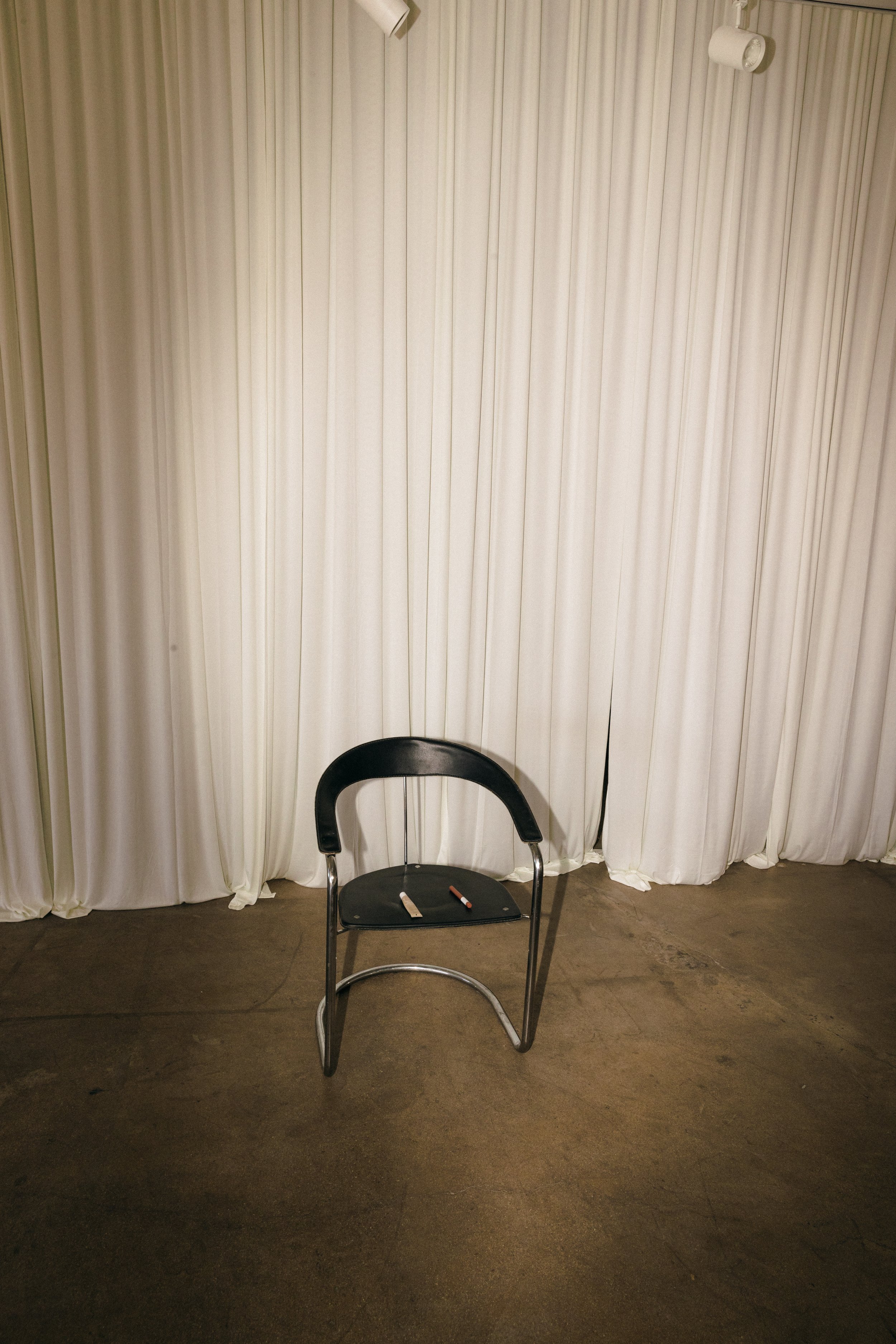 A black chair with a curved backrest on a modern concrete floor, with white curtains in the background and two small objects on the seat.