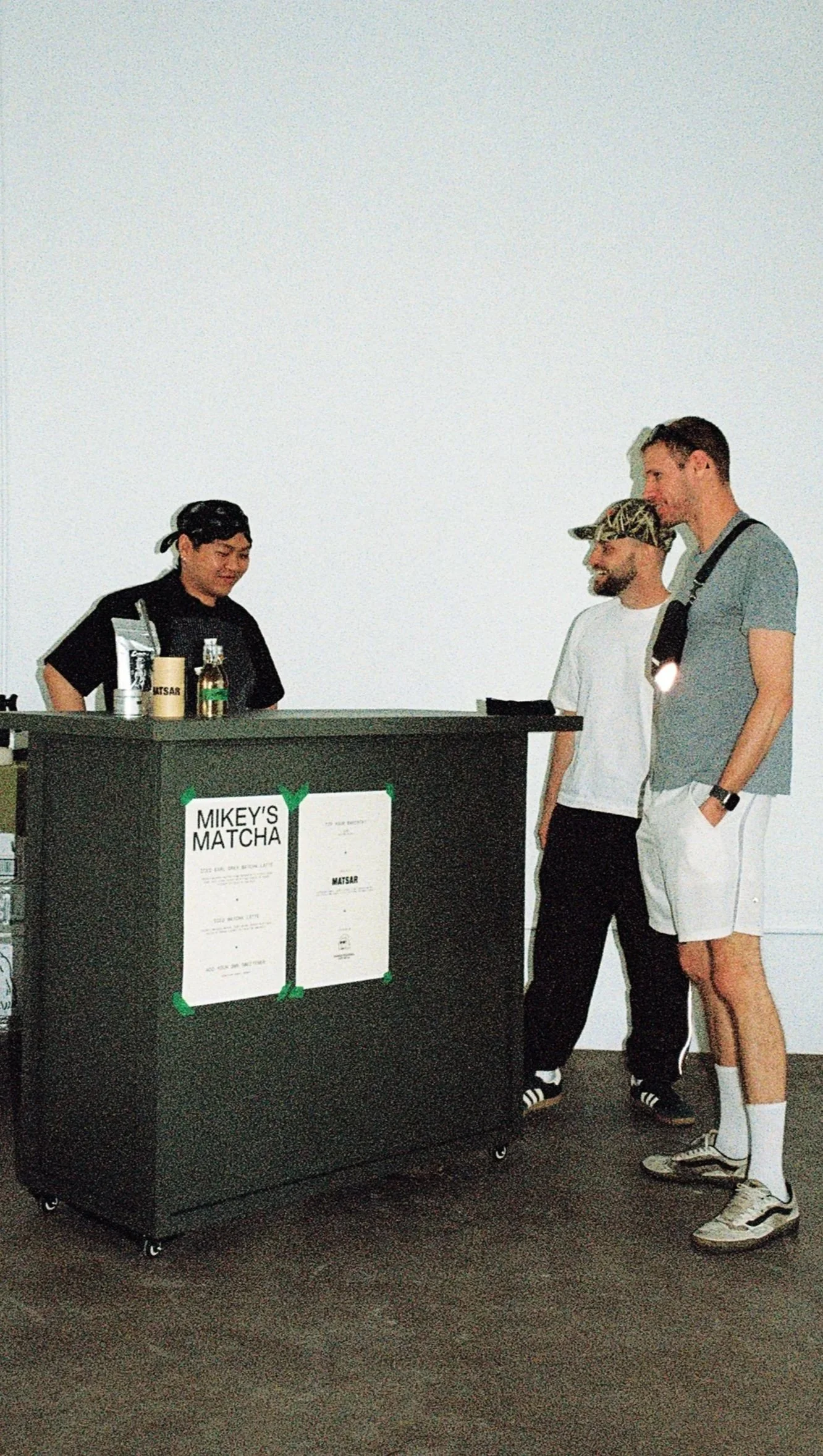 Three men are standing at a black counter with a sign that reads "Mikey's Matcha." One man behind the counter is serving drinks, while the other two are waiting and smiling. The men in line are dressed casually, with one wearing a gray t-shirt and shorts, and the other in a white t-shirt and black pants. The scene appears to be inside a minimalist shop or cafe with a plain white wall.