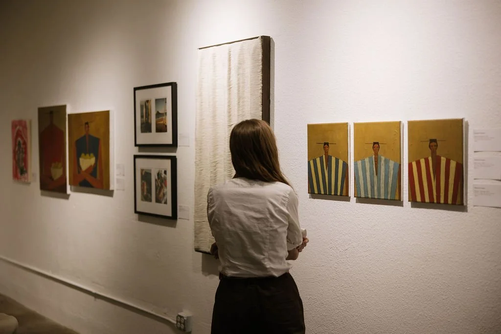 A woman with long brown hair wearing a black tank top, gray jeans, and white sneakers stands in front of a white gallery wall observing framed photographs.