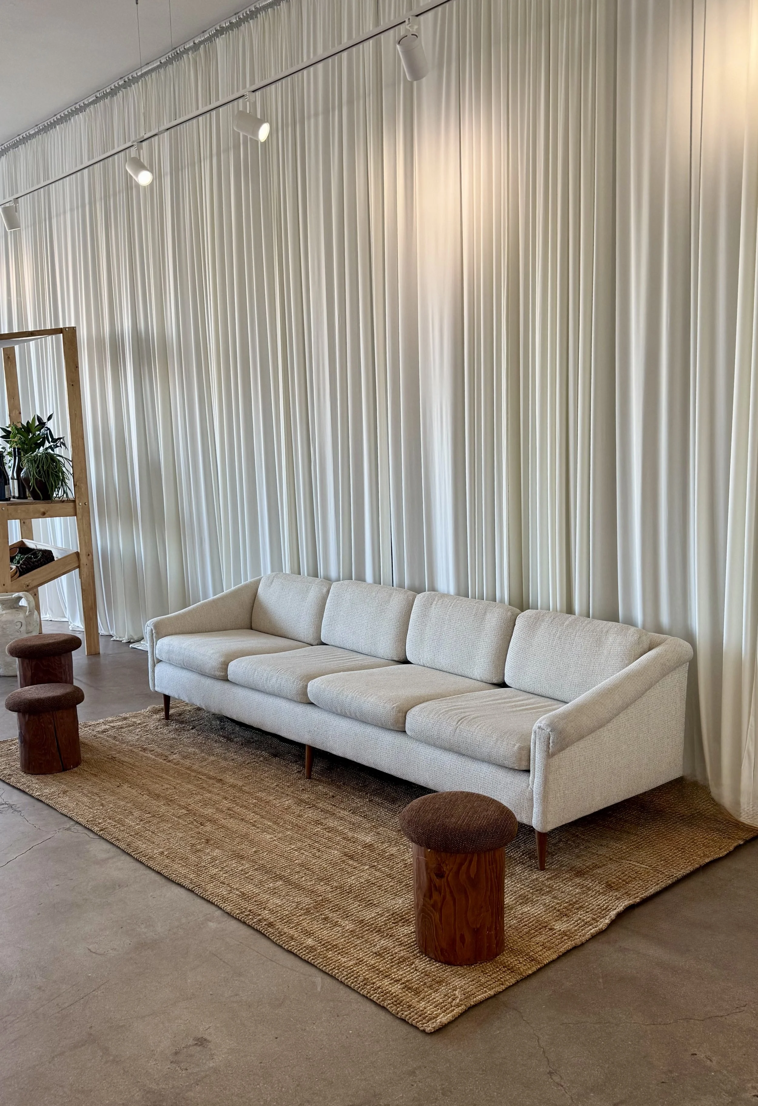 A minimalist interior with a long beige sofa on a woven rug, paired with two small wooden stools, against a backdrop of cream-colored curtains and track lighting.