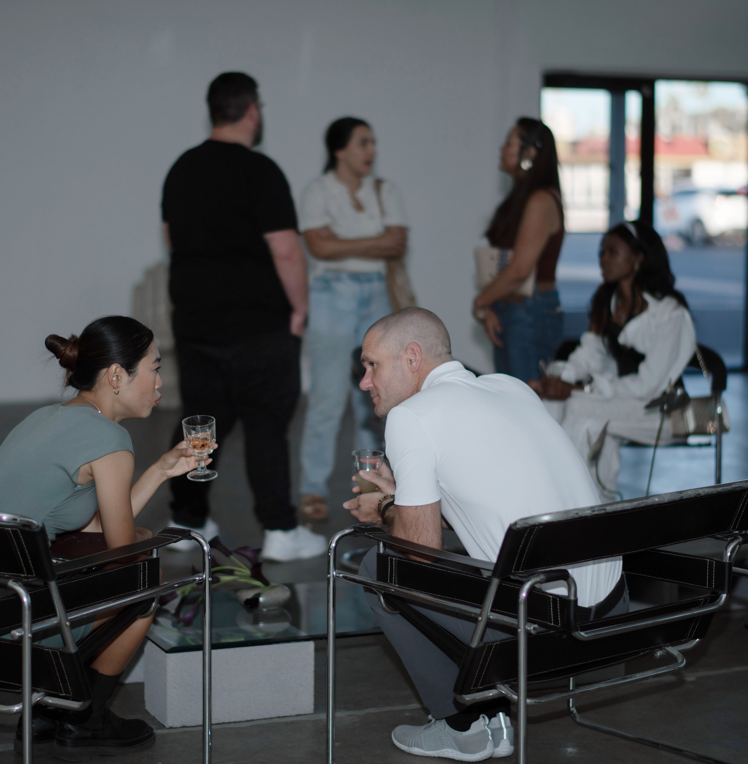 A group of six diverse individuals in a modern indoor space, some sitting and some standing, engaged in conversation, with large windows in the background.