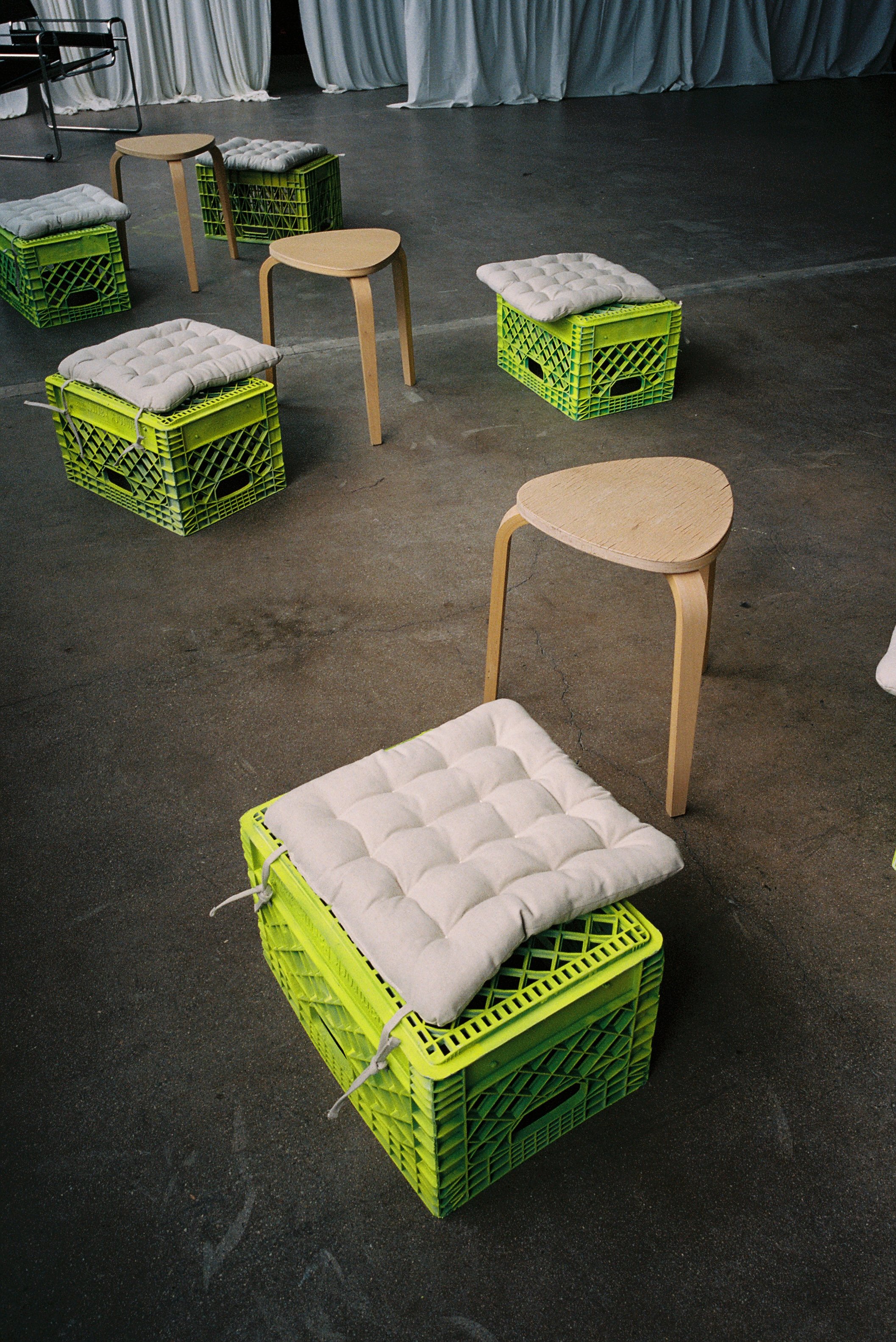 Arrangement of wooden stools and cushions on green plastic crates in a room with curtains.