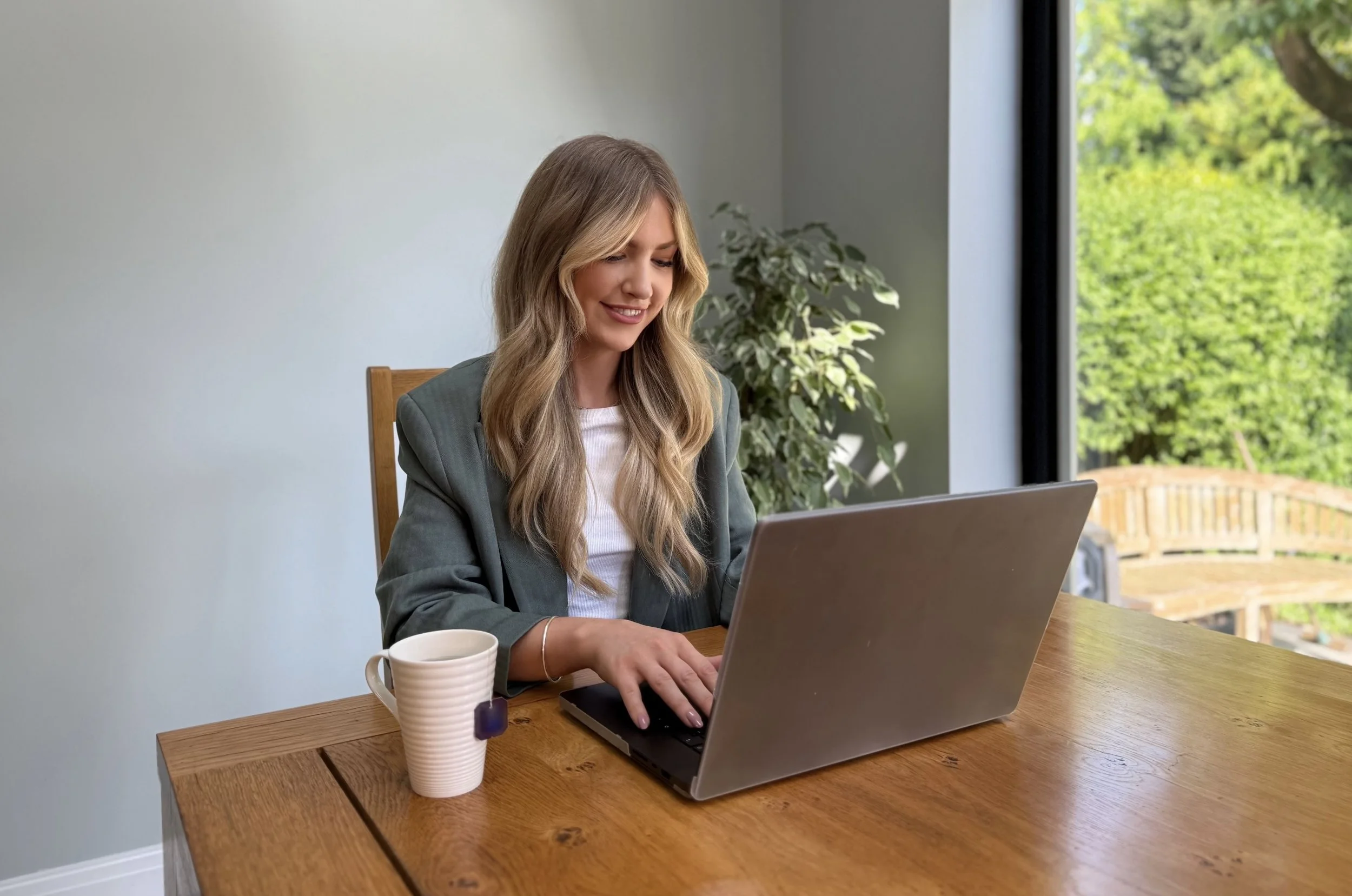 Woman using a laptop at a wooden table with a coffee mug nearby, in a bright room with large window showing greenery outside, representing a nutrition therapy consultation