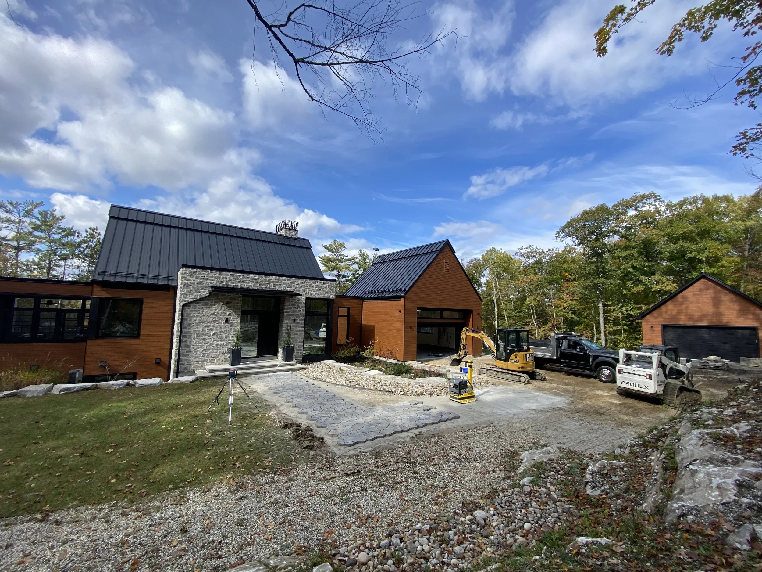 Construction d'une maison moderne avec plusieurs véhicules et machines sur le terrain, entourée d'arbres, sous un ciel bleu avec quelques nuages.