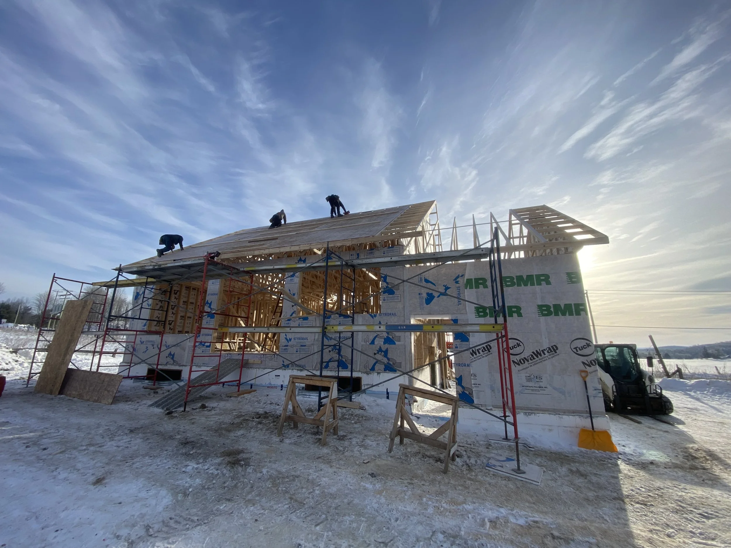 Construction en cours d'une maison en bois sous un ciel bleu avec quelques nuages, dans un environnement enneigé.