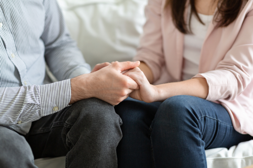 Close-up of a couple holding hands, symbolizing support and comfort, seated on a couch.